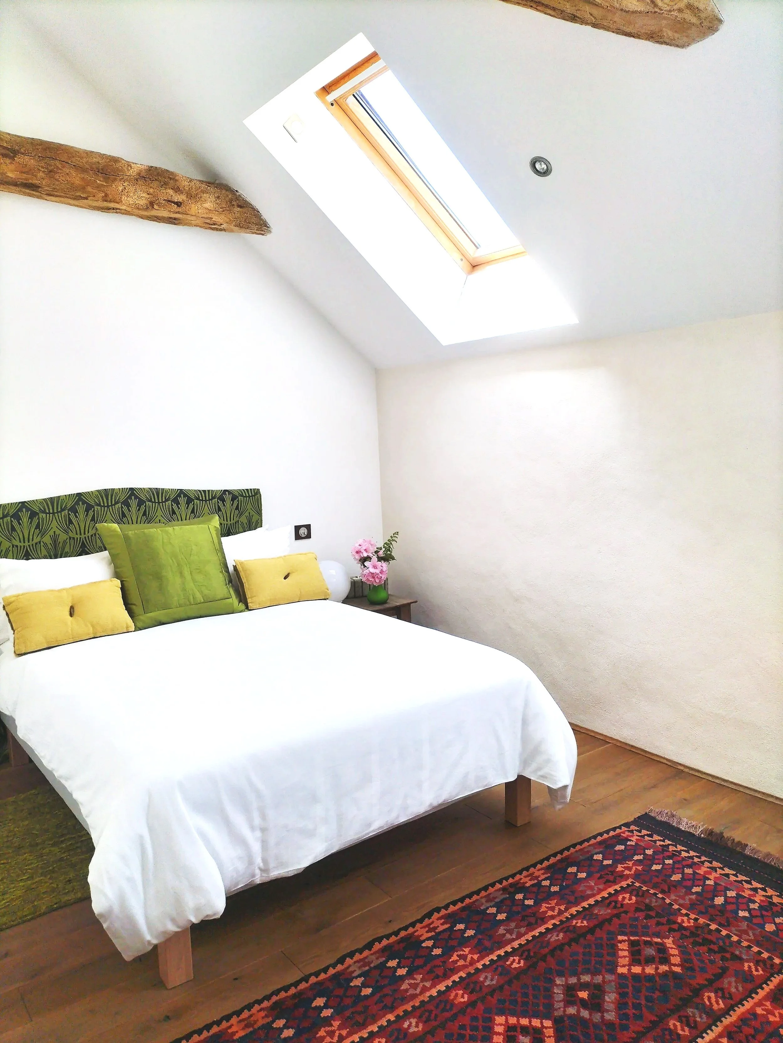 A bedroom with white walls and wooden beams, featuring a bed with white bedding and green and yellow pillows, a bedside table with pink flowers, a red patterned rug, and a skylight window in the ceiling.