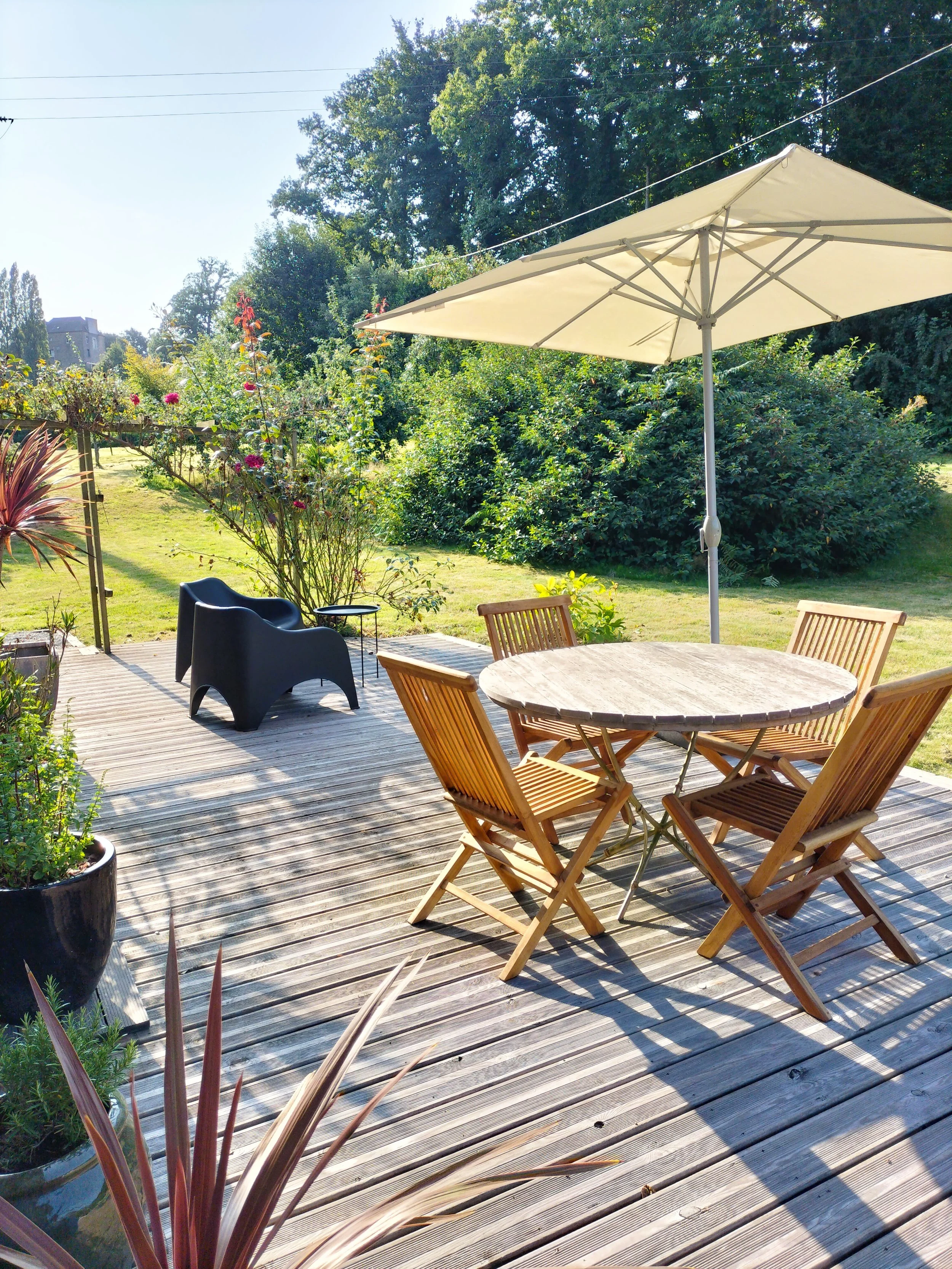 Outdoor wooden deck with a round wooden table, four wooden chairs, a large beige patio umbrella, black outdoor chairs, potted plants, and a green garden with trees and bushes under a clear sky.