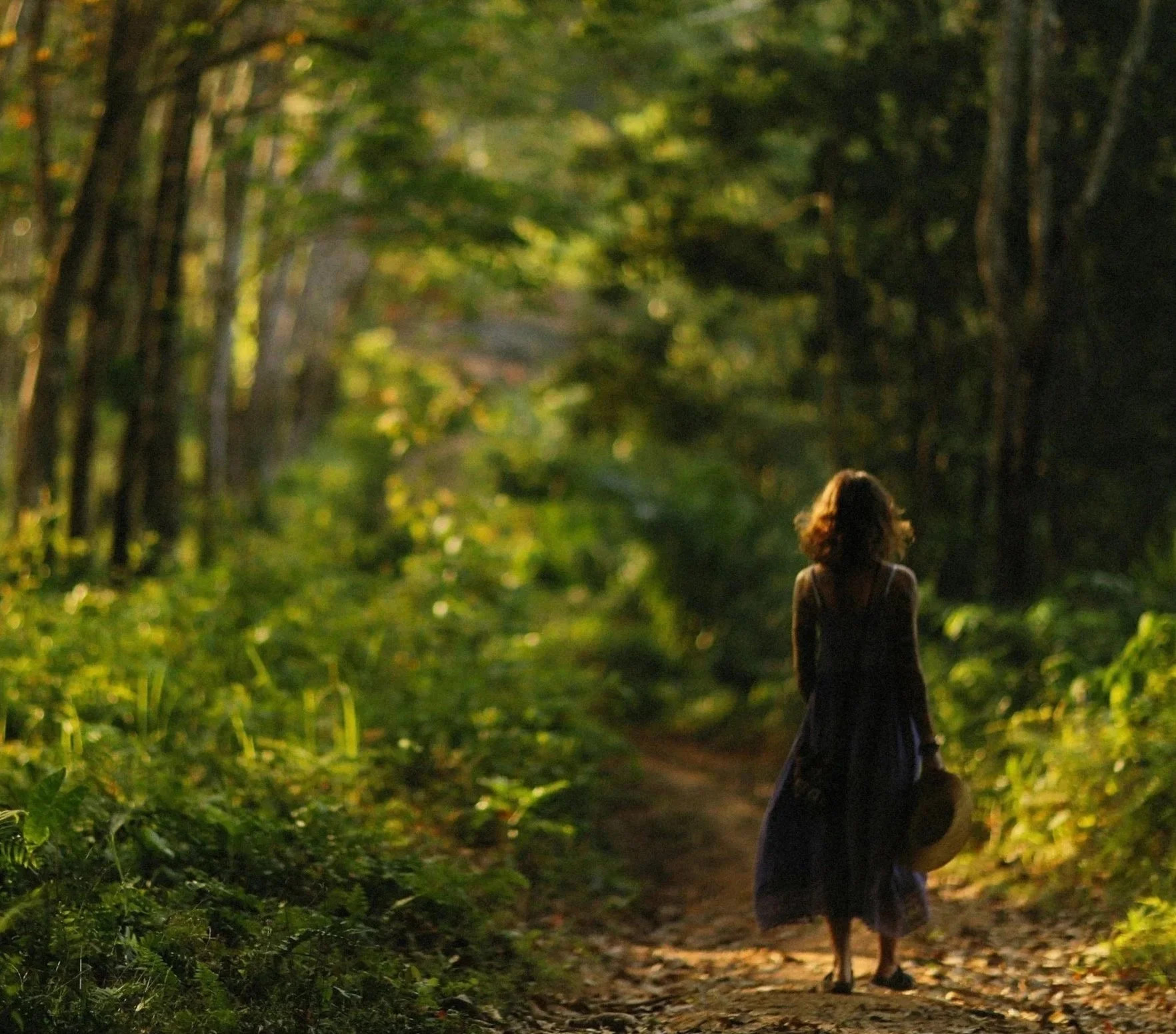A woman walks on a wooded trail during daylight, holding a hat in her hand, surrounded by green trees and foliage.
