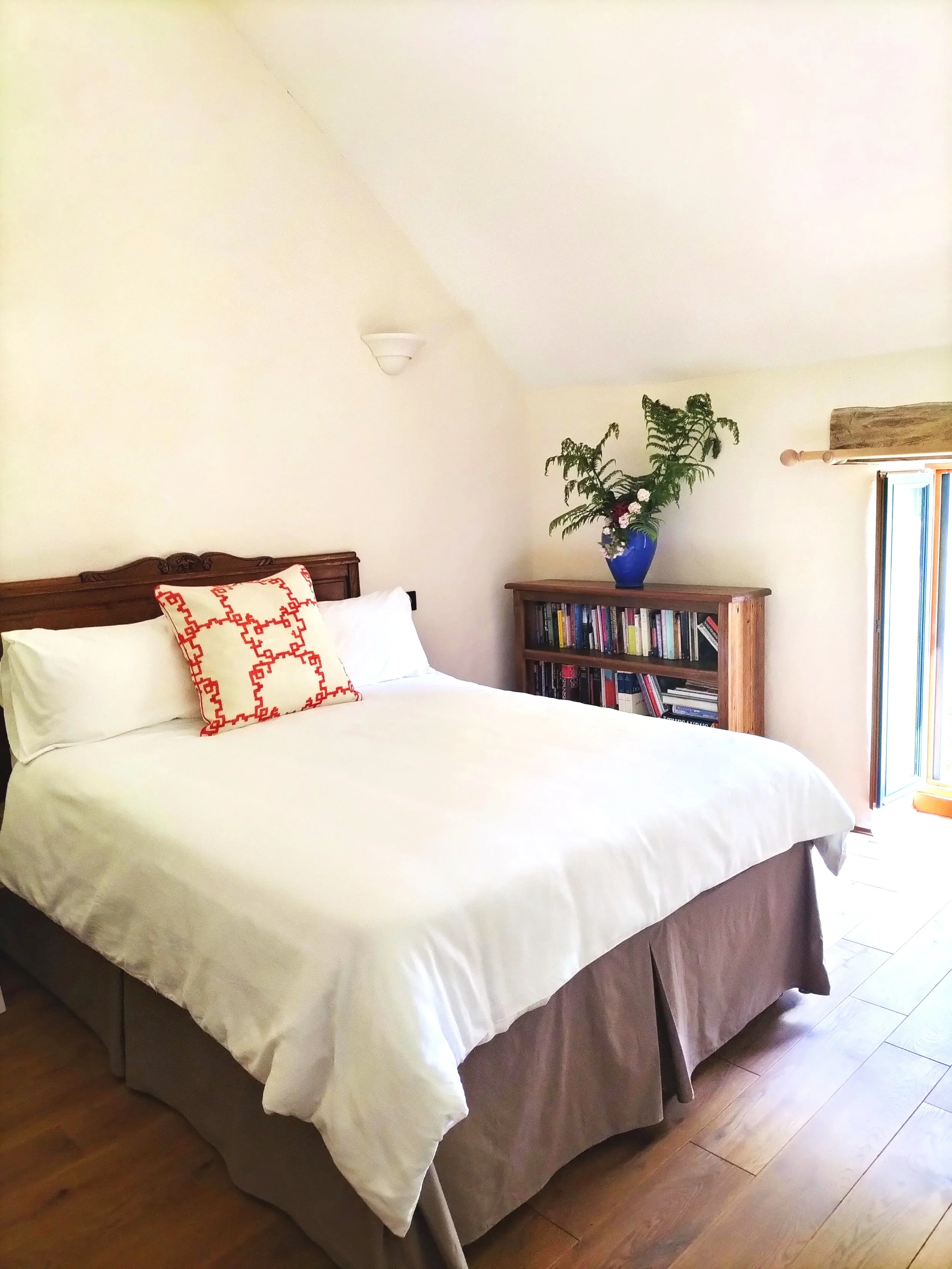 Cozy bedroom with a double bed featuring white linens and a decorative pillow, next to a wooden bookshelf with books and a potted plant.