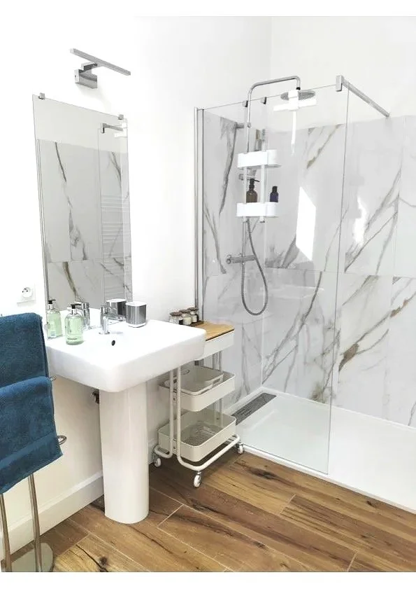 Modern bathroom with wood flooring, white pedestal sink, wall-mounted mirror, walk-in glass shower enclosure with marble-like tiles.