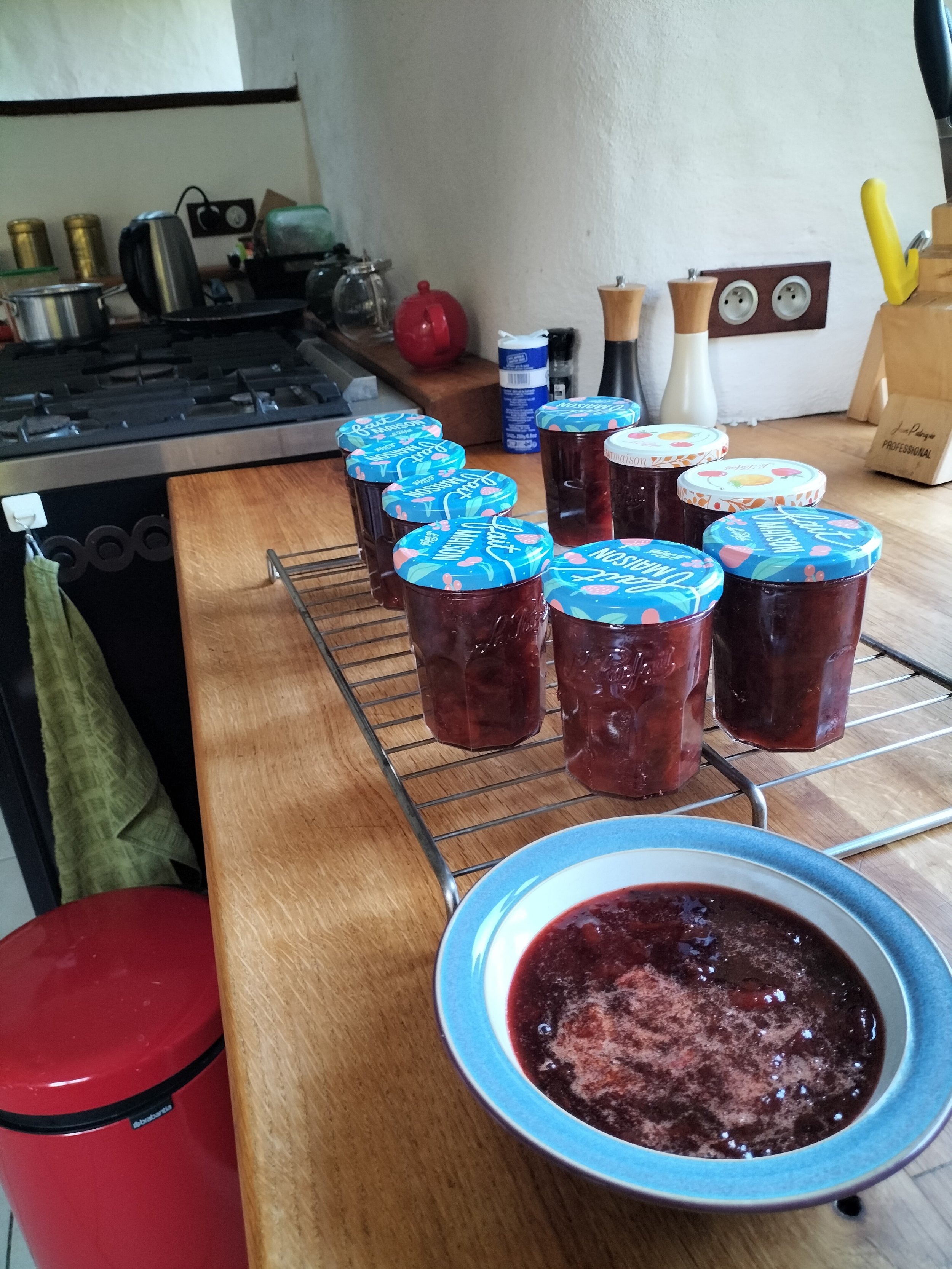 Jars of homemade jam on a kitchen counter next to a bowl with more jam, in a domestic kitchen setting.