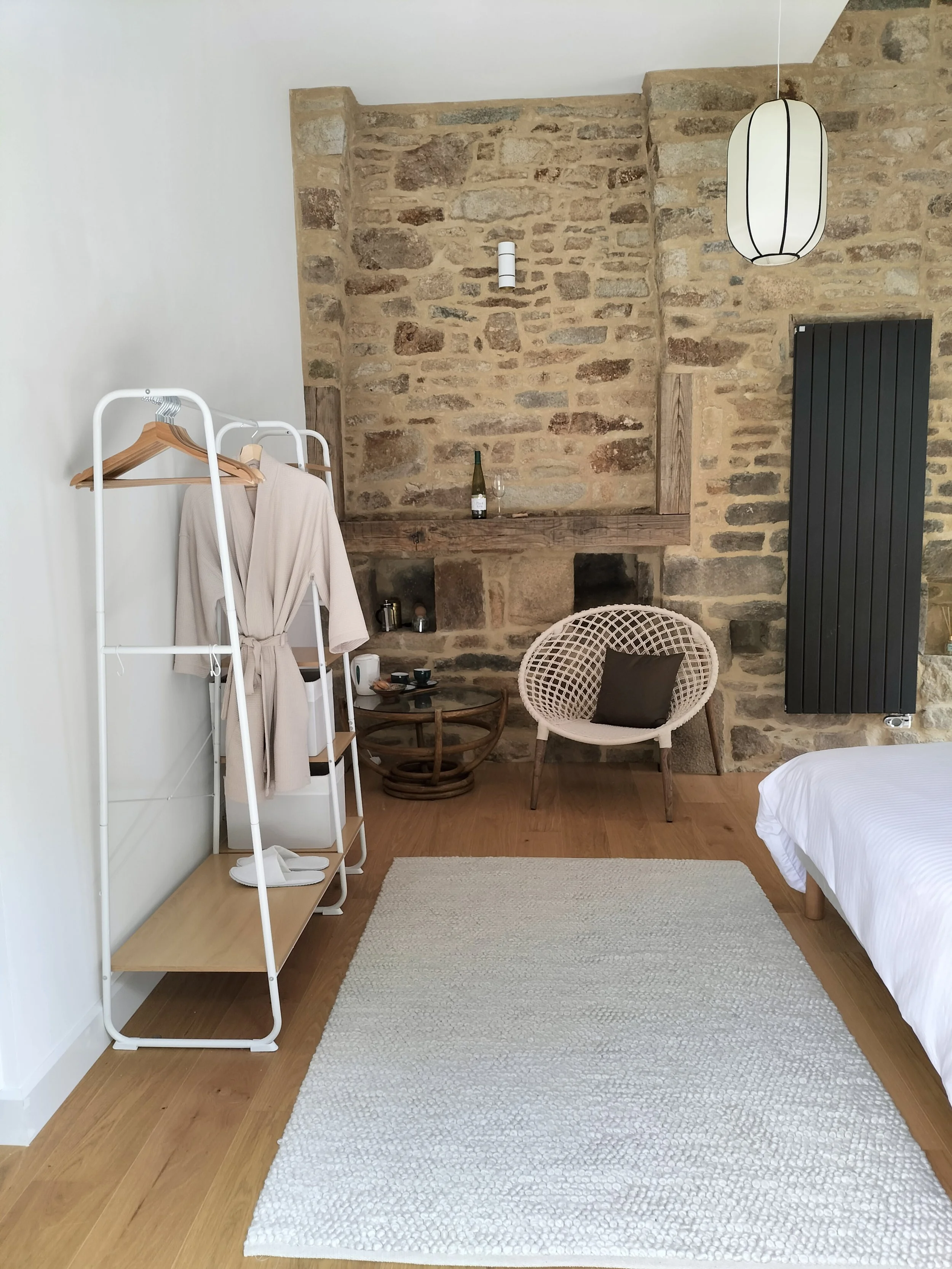 A bedroom with a stone fireplace, a hanging paper lantern, a wicker chair with a pillow, a small table with a bottle and glass, a white clothing rack with robes, a white bed, a black radiator, and a cream-colored rug.