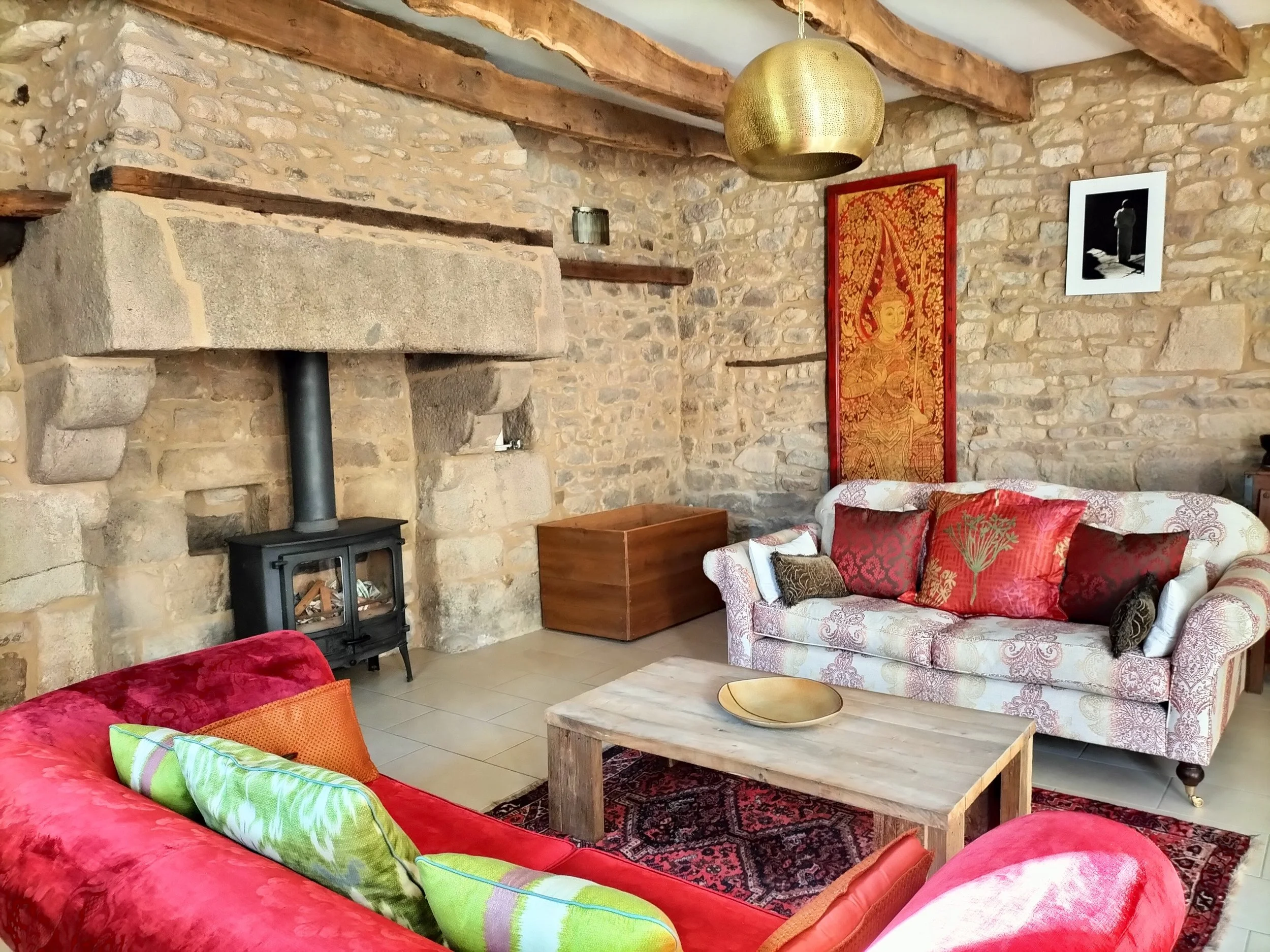 Living room with stone walls, a black wood stove, and wood beams on the ceiling. There are colorful cushions on a floral-patterned sofa, a wooden coffee table, and artwork on the walls including a red and gold tapestry.