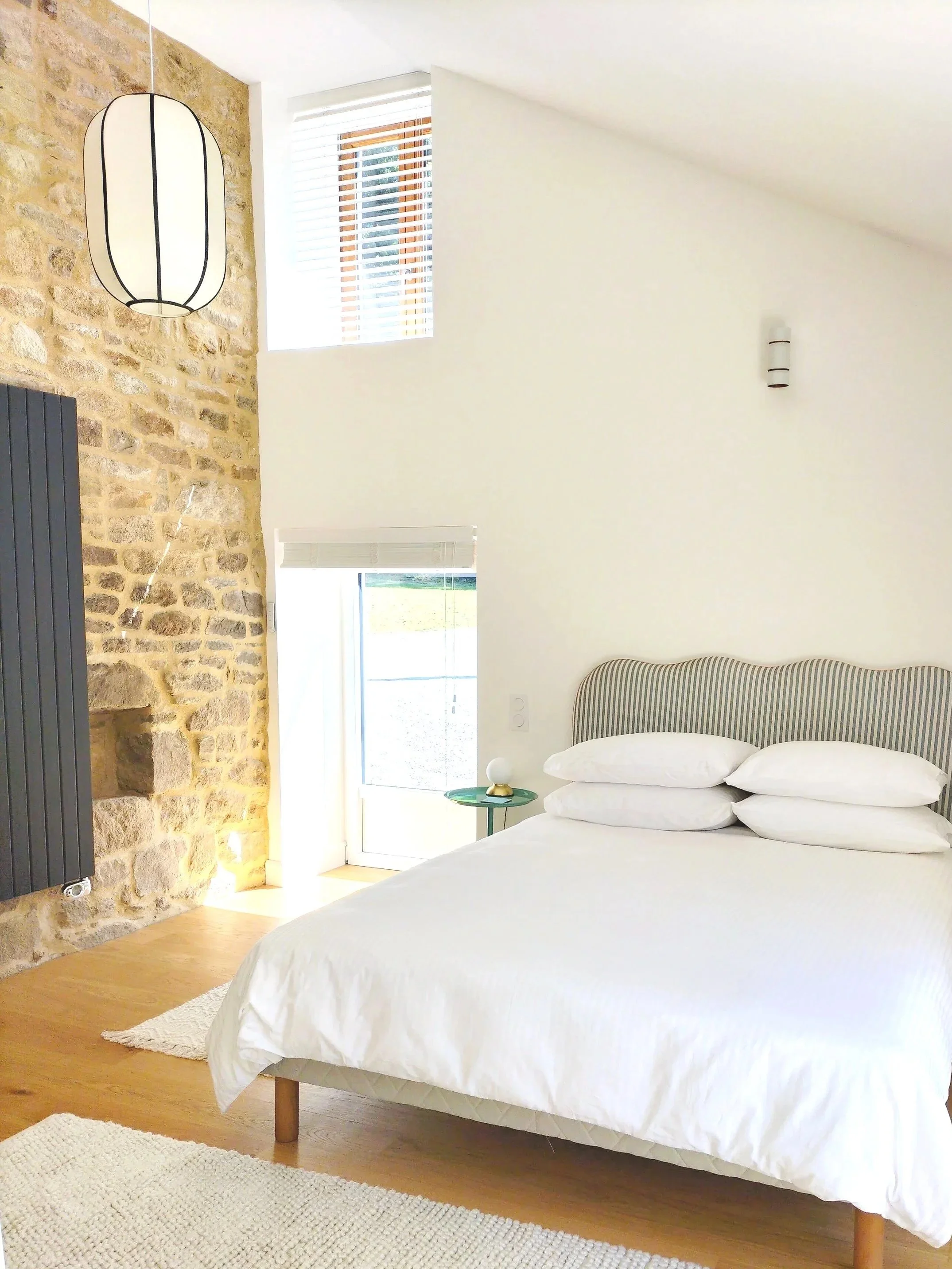 Bright bedroom with a white bed, striped headboard, small green side table, stone accent wall with hanging pendant light, window with blinds, and a door leading outside.