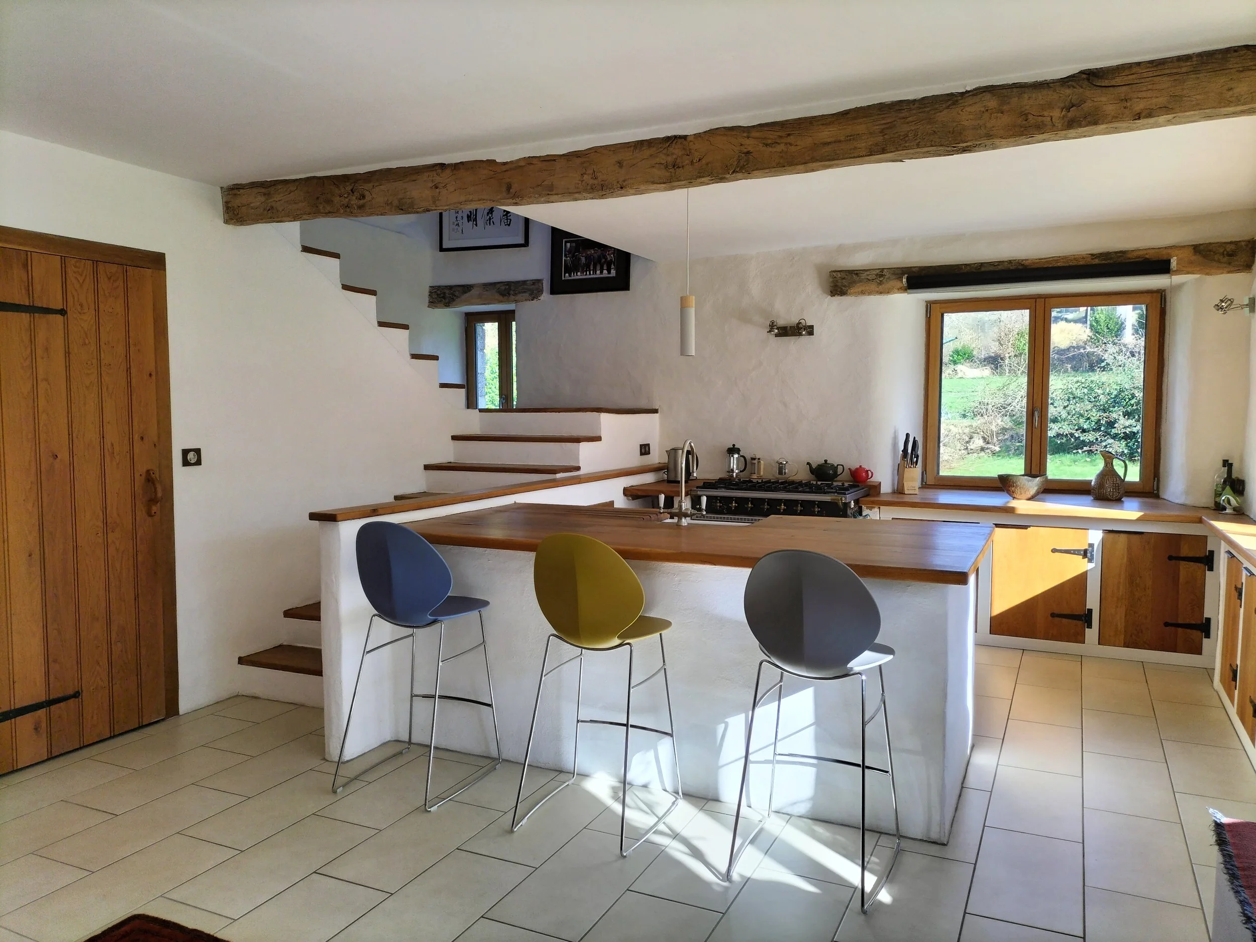 Modern kitchen with wooden countertops, white textured walls, a large window showing greenery outside, and three colorful bar stools in blue, yellow, and black in front of the island.