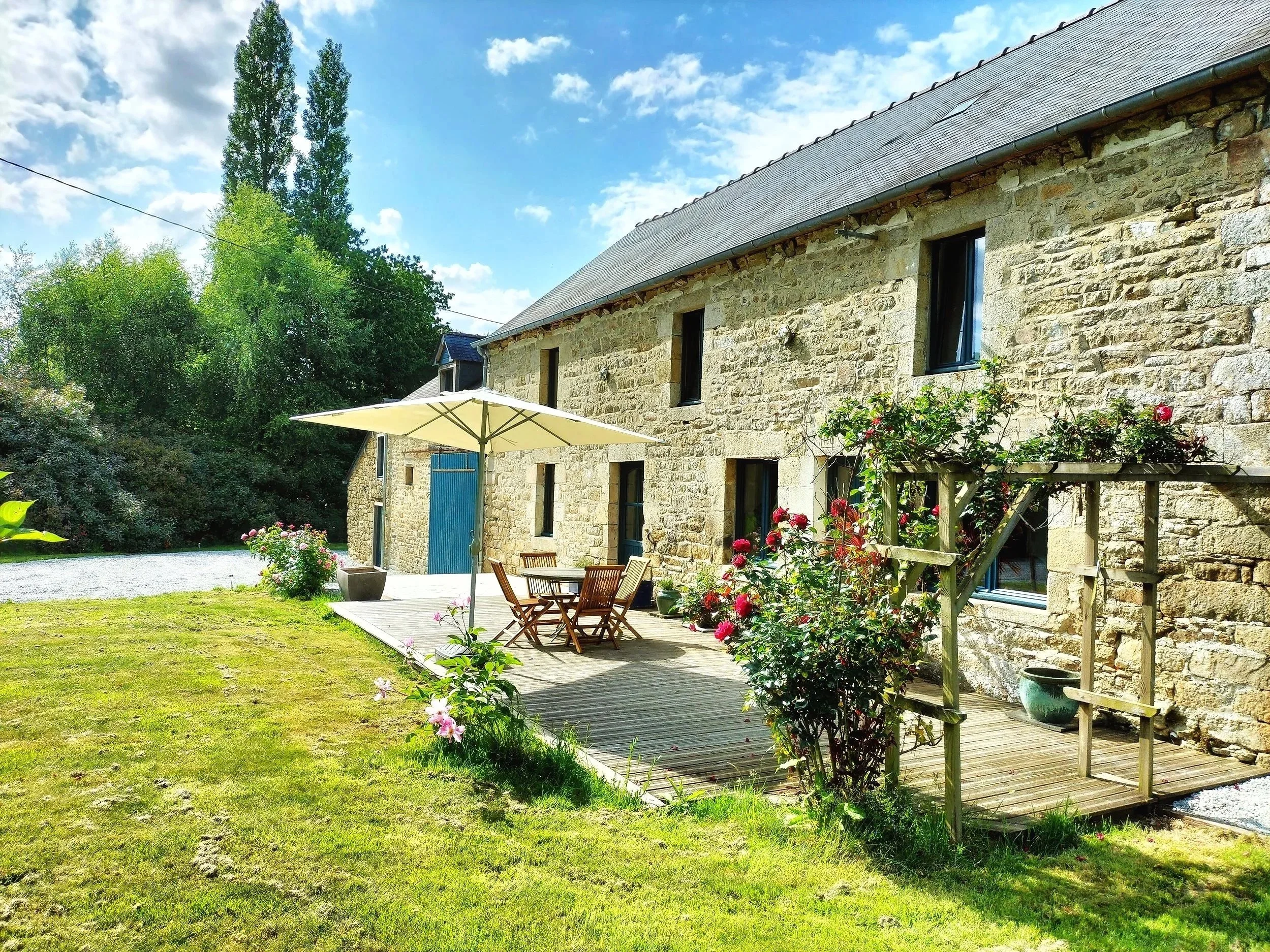 Stone house with a wooden deck and outdoor seating area, surrounded by a grassy yard and blooming flowers under a partly cloudy sky.