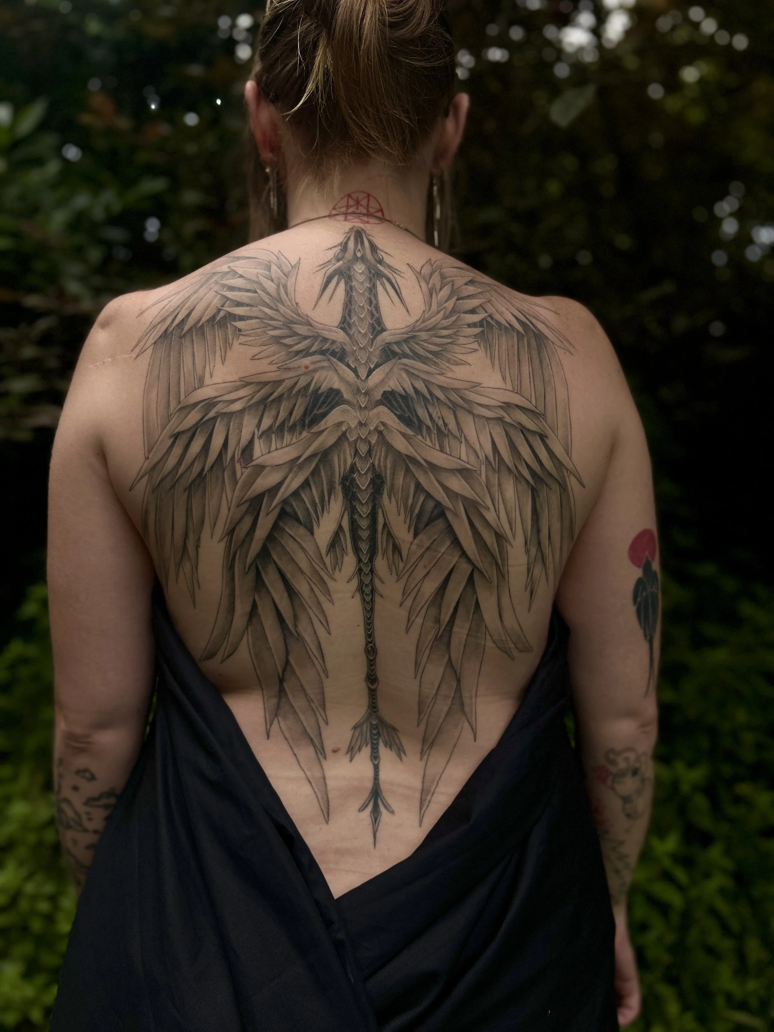 Tatouage d'un dragon déployant ses ailes sur le dos d'une femme vue de dos, en pleine nature.
