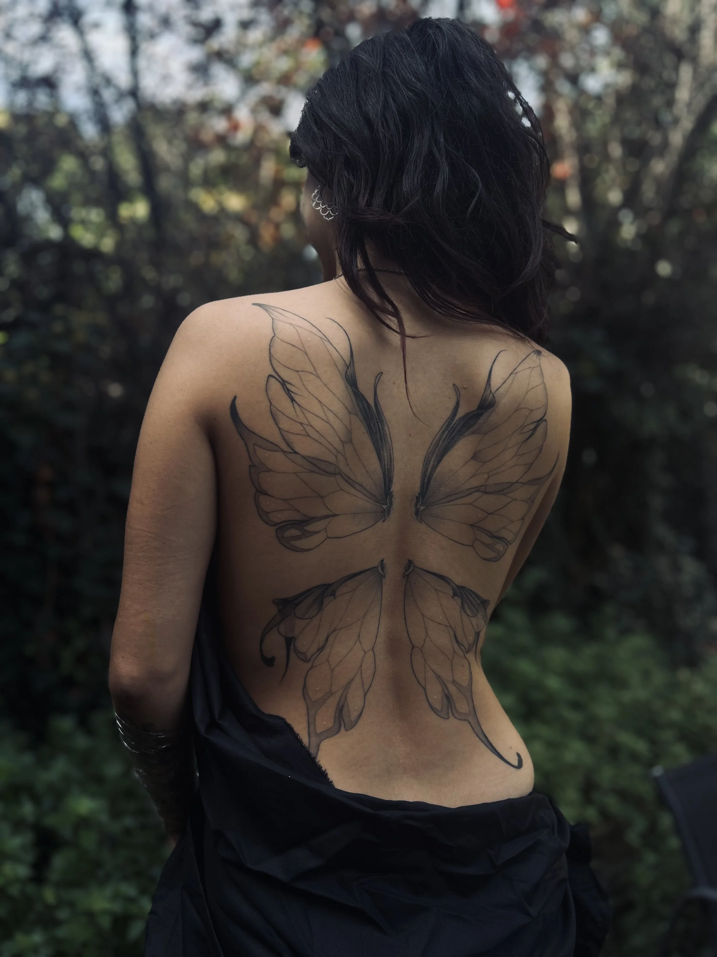 Une personne vue de dos avec un tatouage de papillons sur le dos, dans un environnement naturel avec des arbres en arrière-plan.