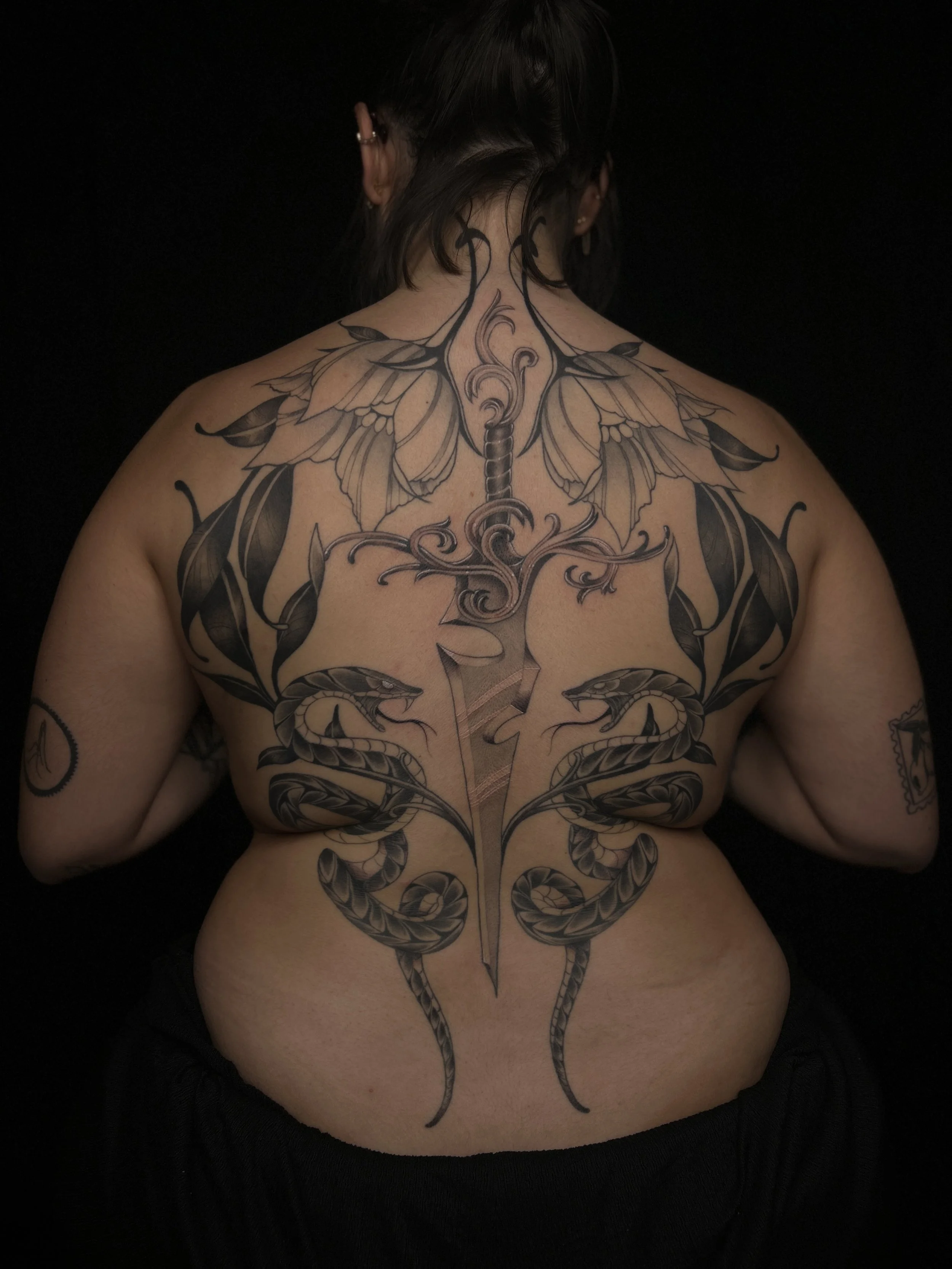 Tatouage complexe sur le dos d'une femme, représentant un arbre avec des branches, des serpents enroulés et des éléments floraux, sur un fond noir.