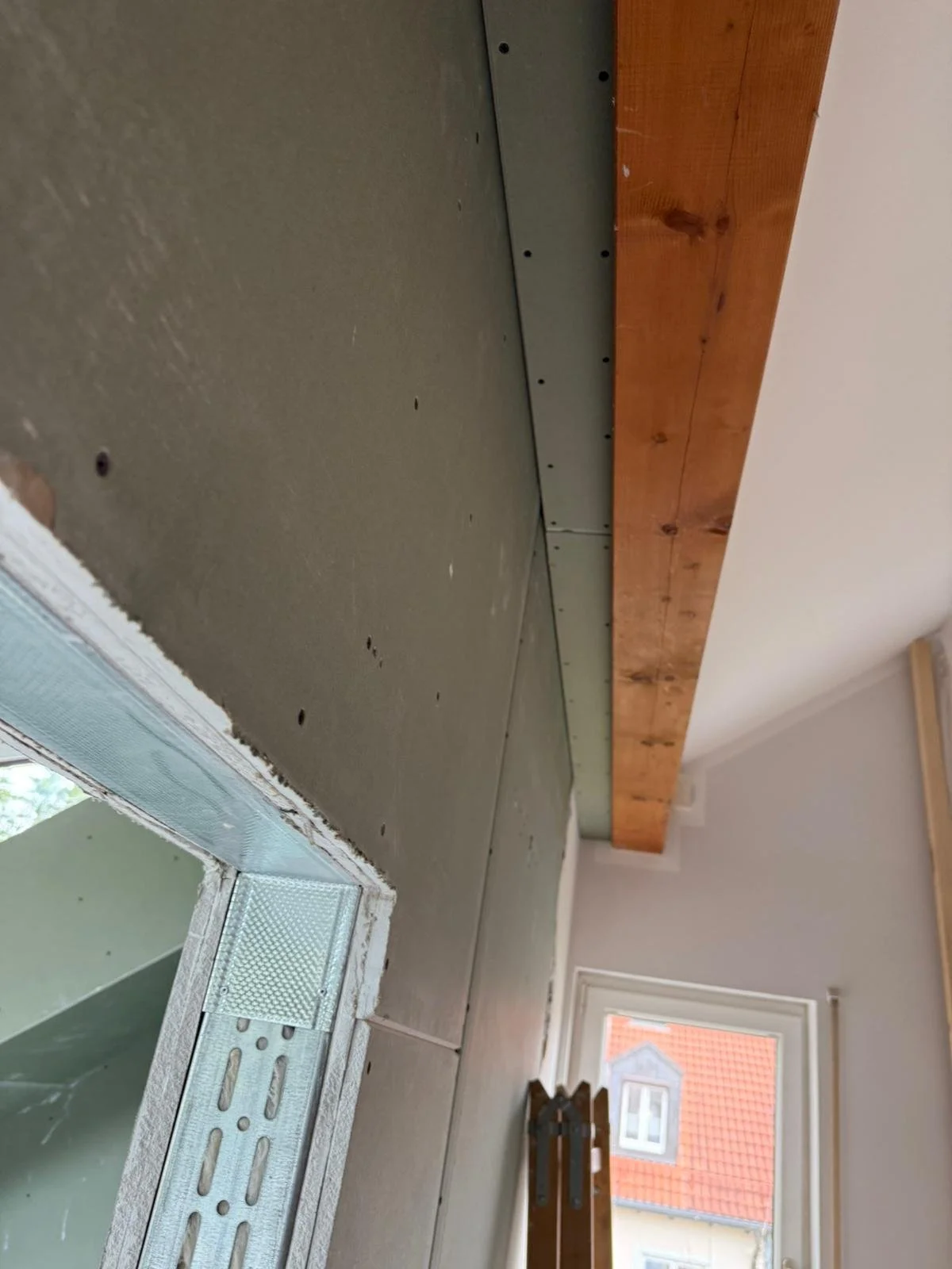 Innenaufnahme eines Raumes während der Renovierung, Fokus auf vorbereiteten Wänden mit Trockenbauplatten, Fenster mit Blick auf Nachbarhäuser.