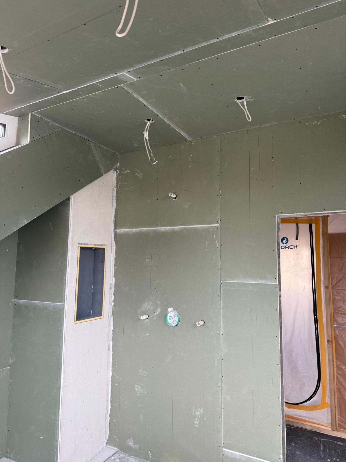 Innenraum eines Hauses im Bauzustand mit noch nicht fertiggestellten Wänden und Decken, grüne Trockenbauplatten, Kabel und Rohrleitungen sichtbar, eine kleine Fensteröffnung und eine Türöffnung mit Schutzfolie.