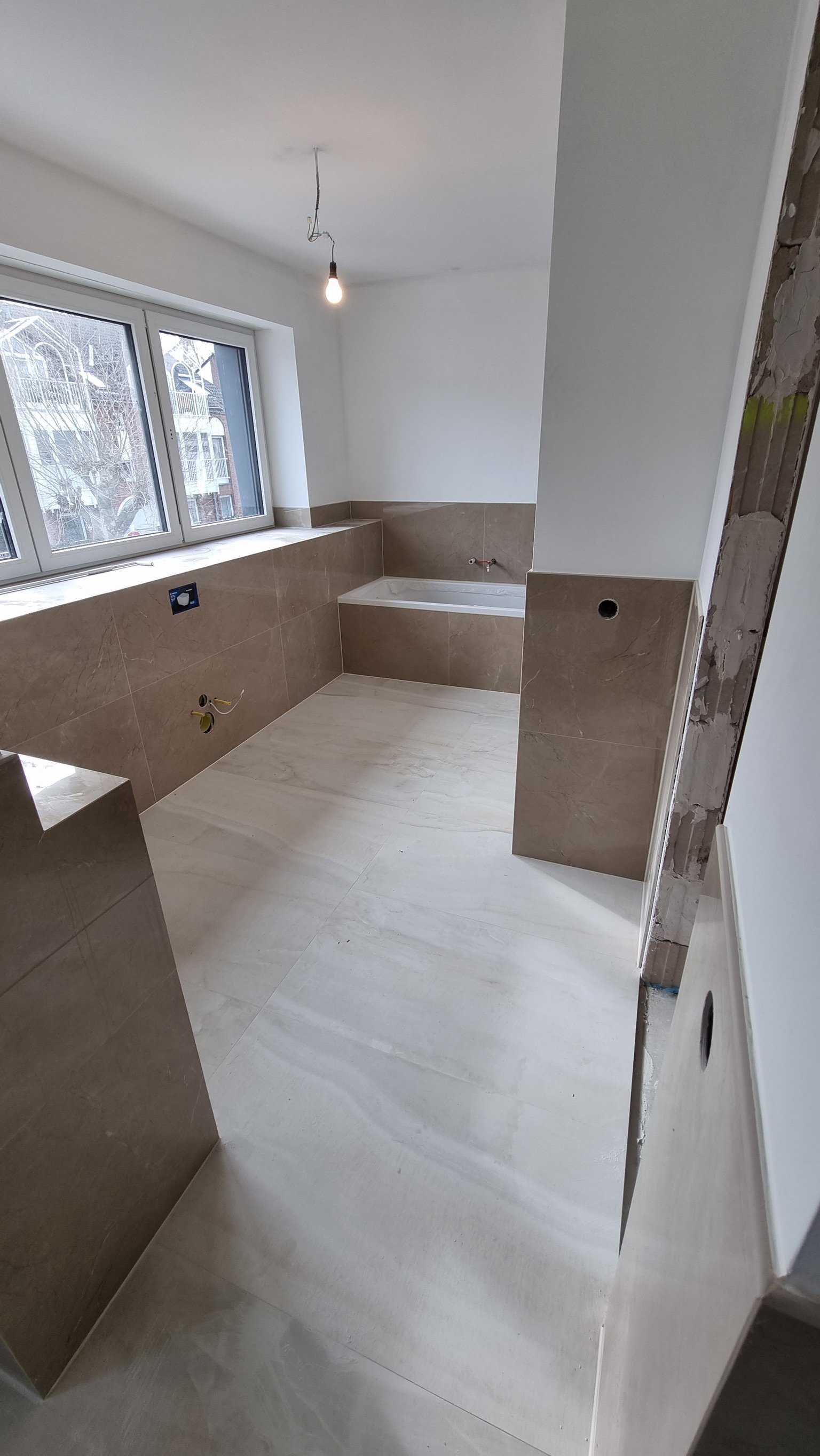 Ein unfertiges Badezimmer mit braunen Wand- und Bodenfliesen, eine große Fensterfront, und ein eingelassener Waschtisch oder Badewanne. Es gibt unvollständige Installationen für Wasser- und Stromanschlüsse.