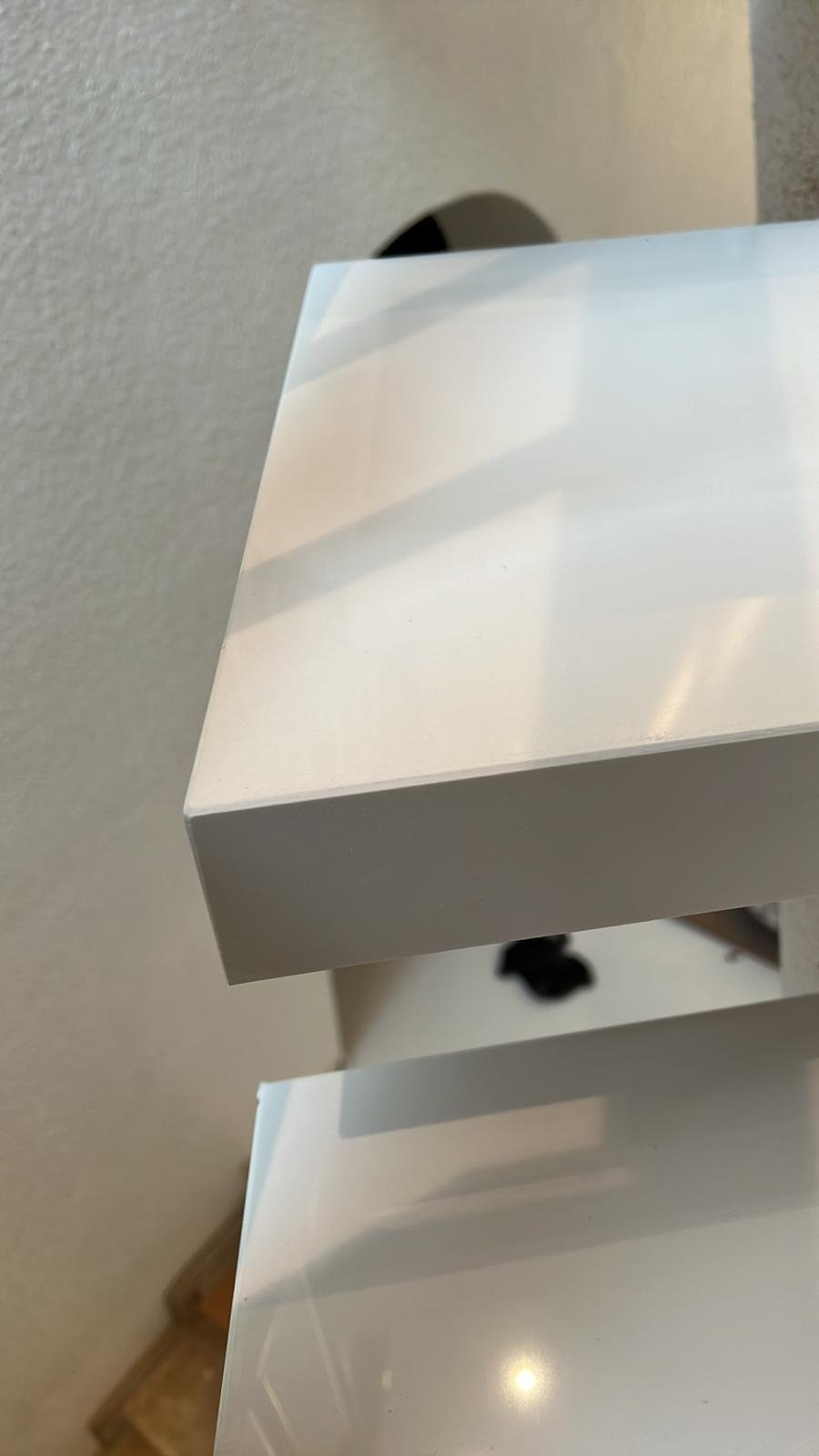 Nahaufnahme eines weißen, glänzenden Objekts, wahrscheinlich eine Verpackung oder Schachtel, mit Schatten auf der Oberfläche, vor einer grauen Wand.