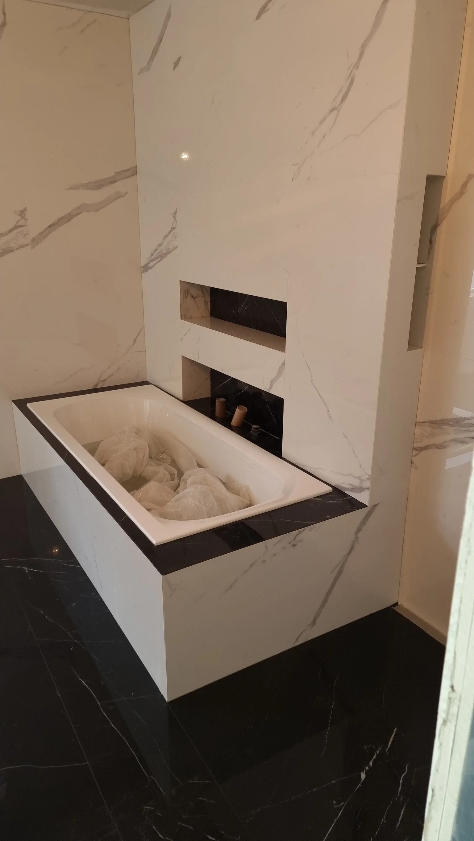 Ein Badezimmer mit schwarzem Marmorboden, weißer Marmorwand und einer eingelassenen Badewanne, die noch verpacktes Material enthält, sowie eine Wandnische für eine Dusche oder einen Kamin.