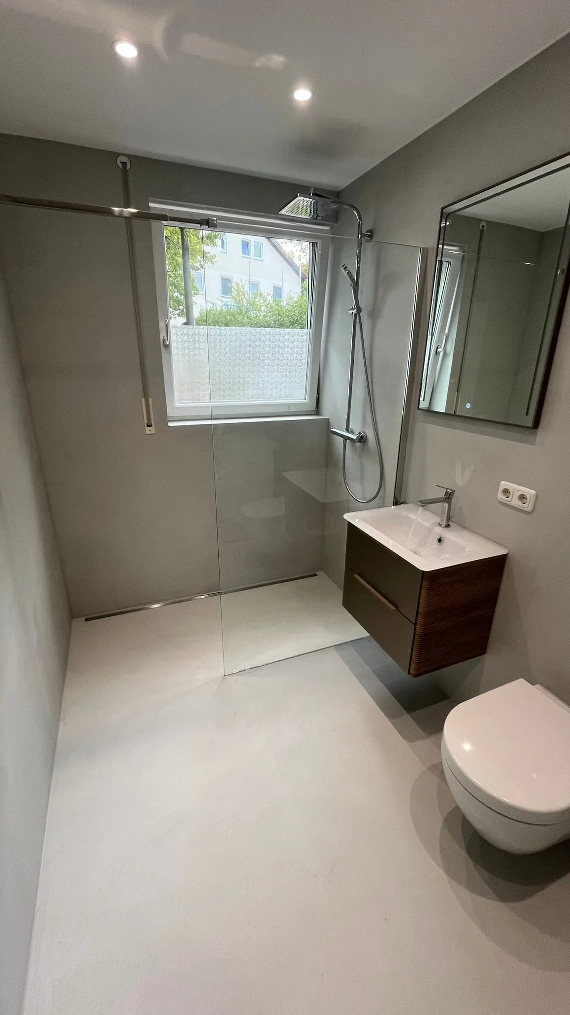 Ein modernes Badezimmer mit einer ebenerdigen Dusche, einem kleinen Waschbecken mit Unterschrank, einem Wandspiegel, einer Toilette und einem Fenster mit Blick nach draußen.