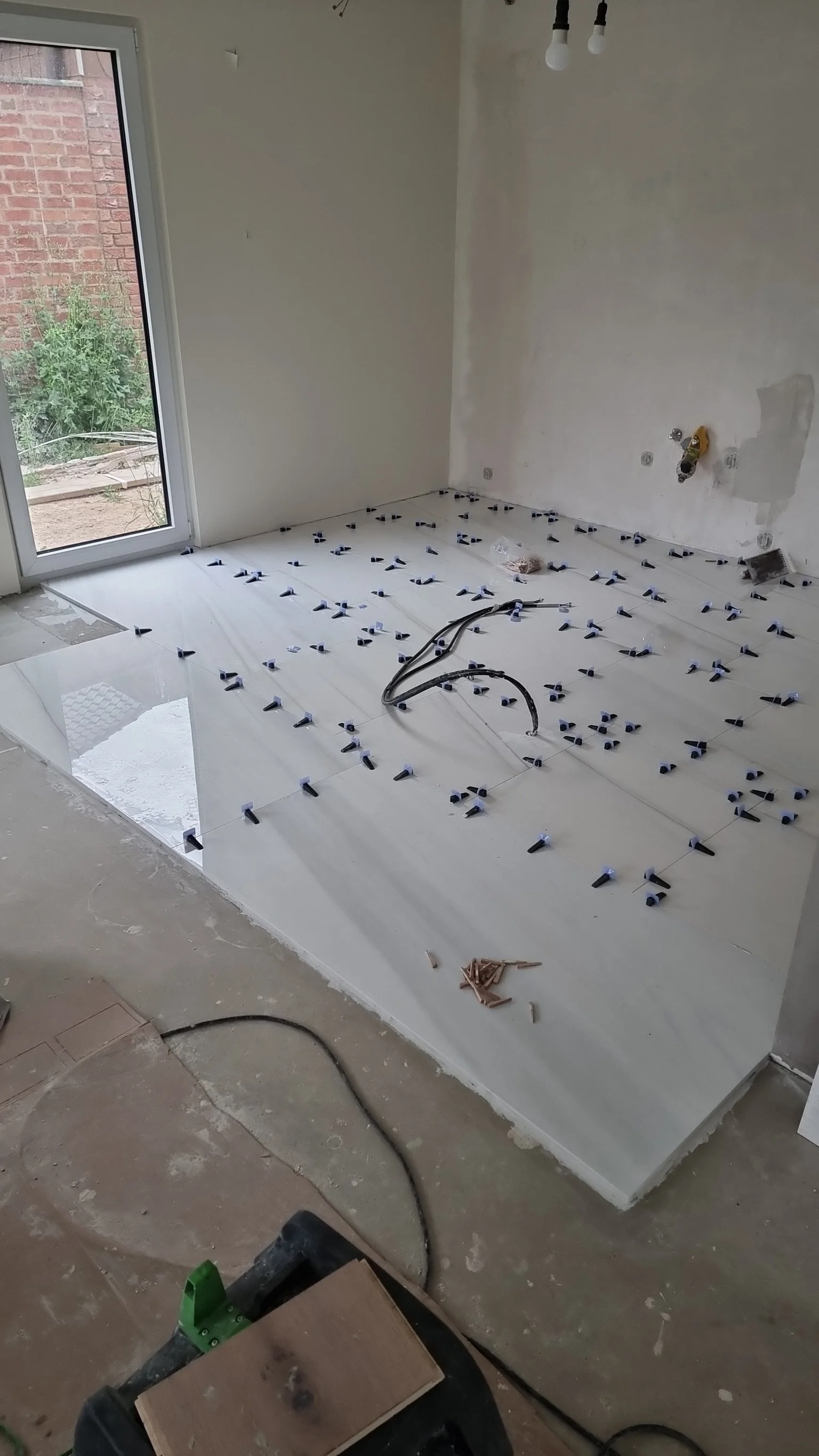 Innenaufnahme eines Raums während der Fußbodenheizung Installationsarbeiten, mit Fliesen und schwarzen Klebebändern auf dem Boden, einer gerade fertiggestellten Terrasse links und unvollständiger Wandgestaltung.