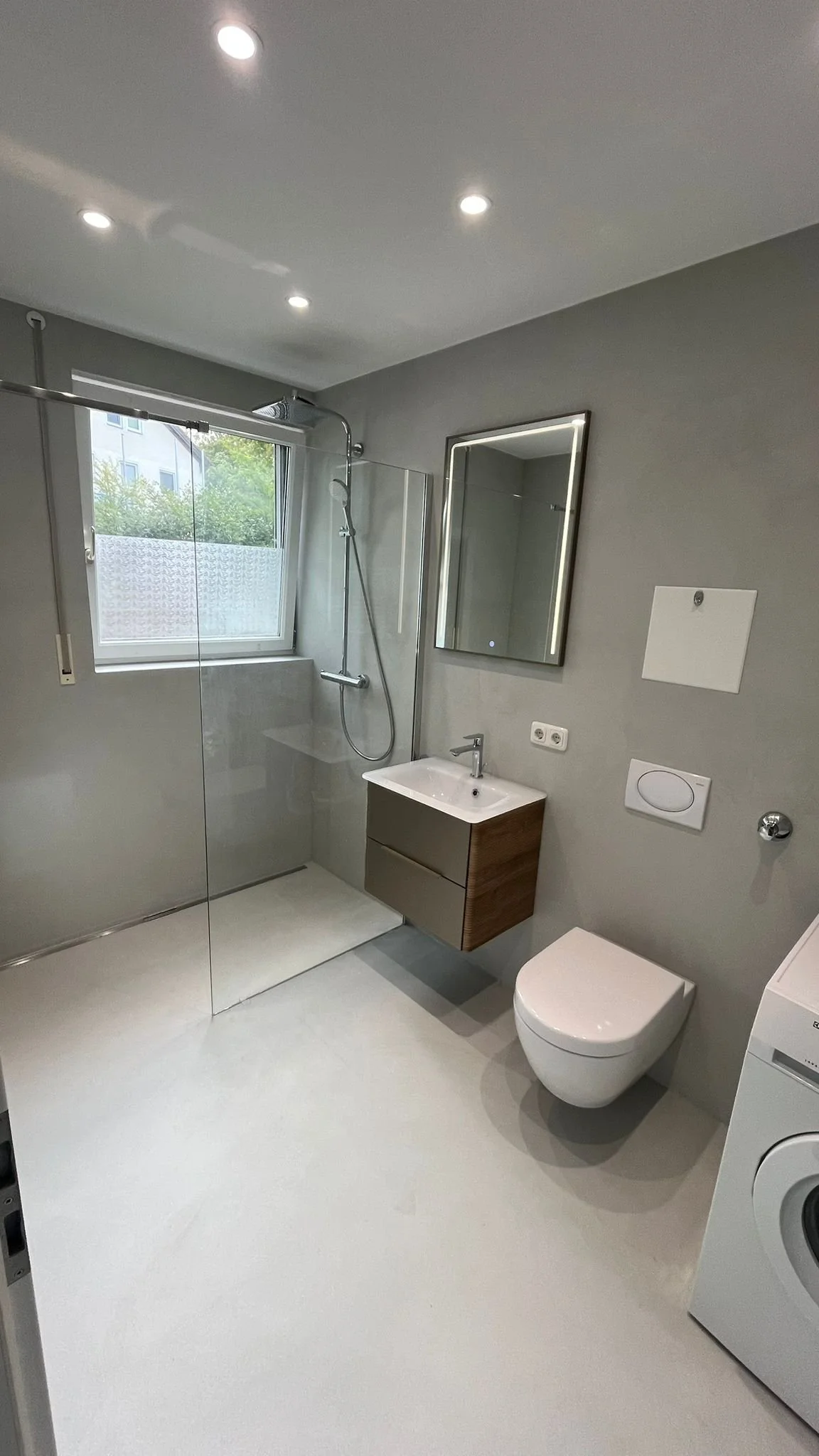 Modernes Badezimmer mit Dusche, Wand-Waschbecken, Spiegel, Toilette und Waschmaschine, helle Wand- und Bodenfarbe, Fenster mit Blick nach außen.