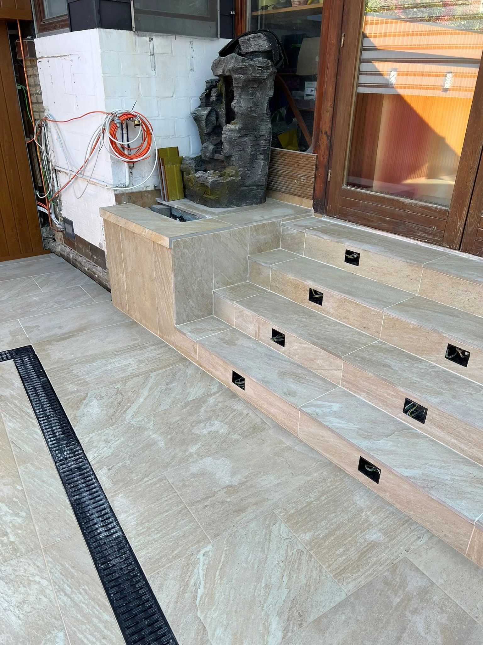 Baustelle mit gefliesten Stufen und Wasserfall-Dekor vor einem Fenster, Arbeiten im Außenbereich, Kabel und Rohre sichtbar.