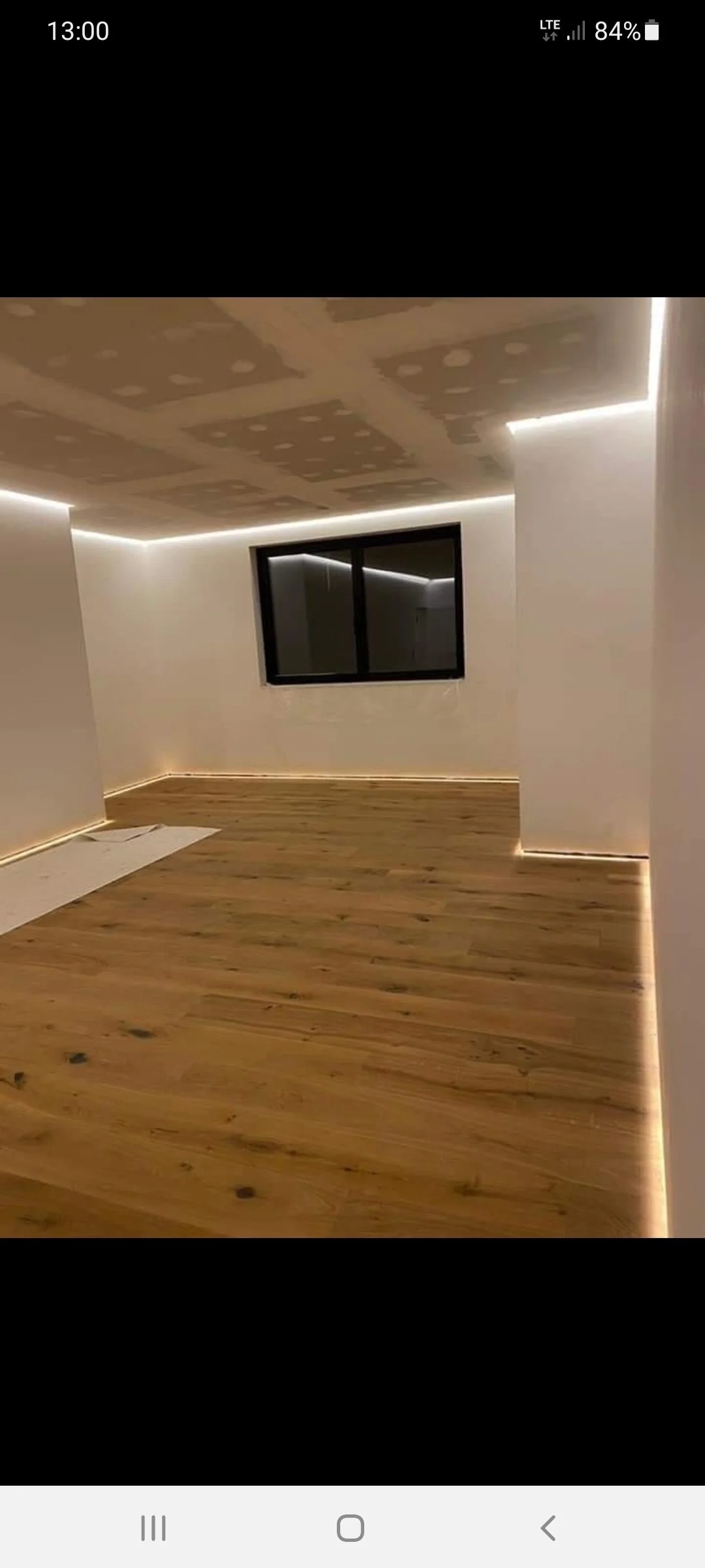 Ein renoviertes Zimmer mit Holzfußboden, weißen Wänden und einem Fenster. Die Decke ist neu verputzt und hat eine LED-Beleuchtung an den Rändern.