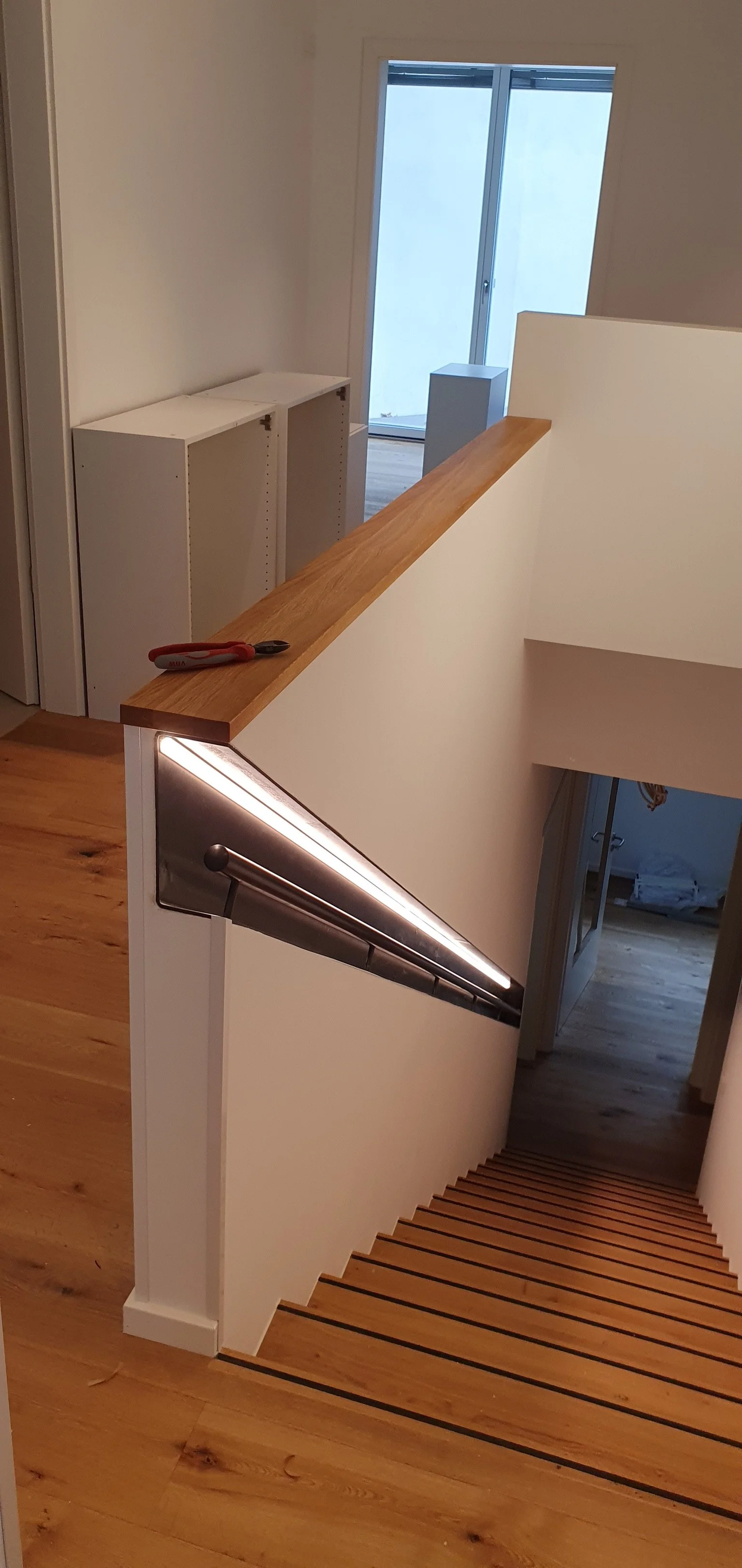 Ein Holztreppenabsatz mit weißen Wänden, Holzgeländer und eingebauter Beleuchtung, Blick nach unten. Im Hintergrund eine Glastür, die in einen Balkon oder einen Außenbereich führt, sowie leere weiße Regale.