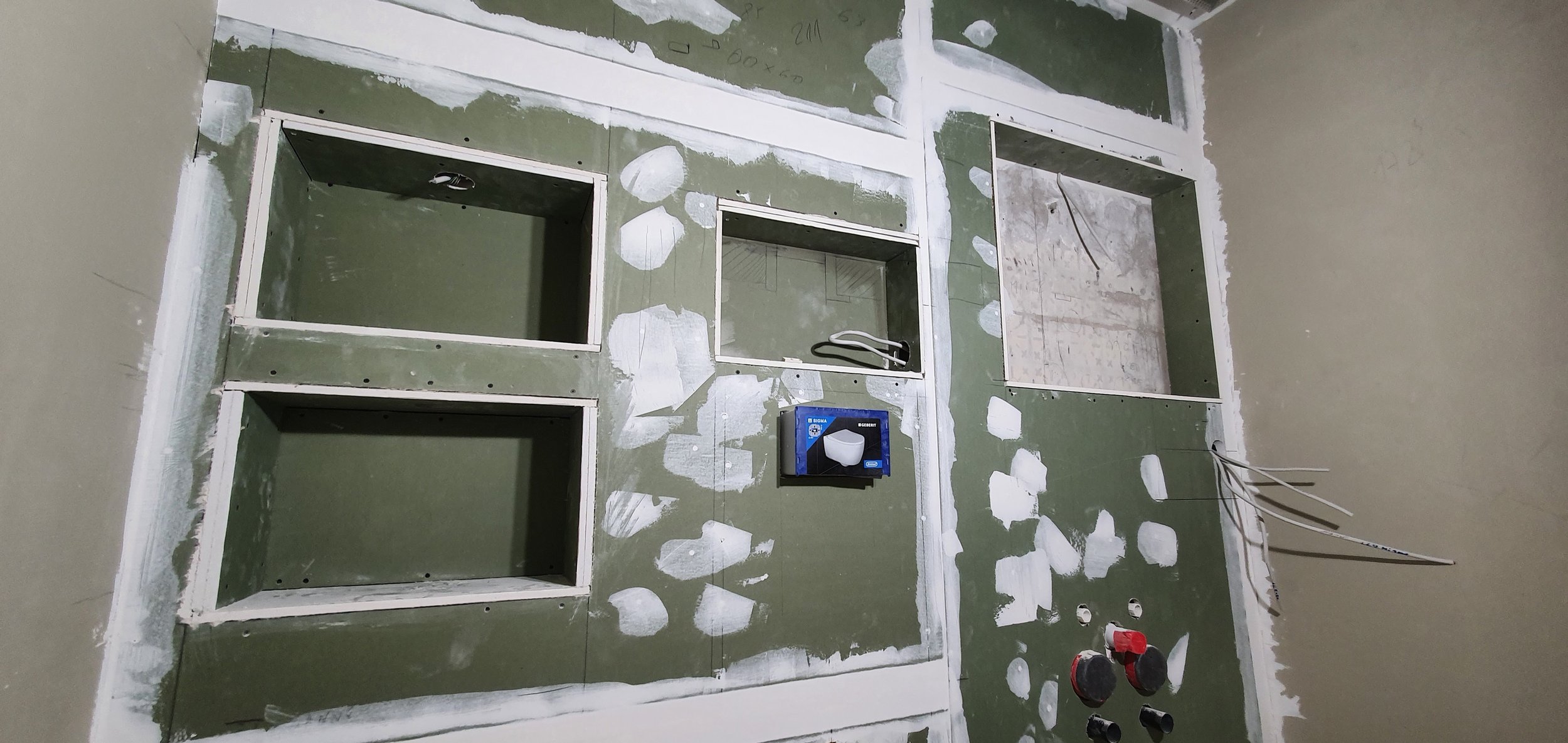 Innenansicht einer Baustelle mit grün kariertem Trockenbau, mehreren Aussparungen, weißen Spachtelstellen und Kabeln.