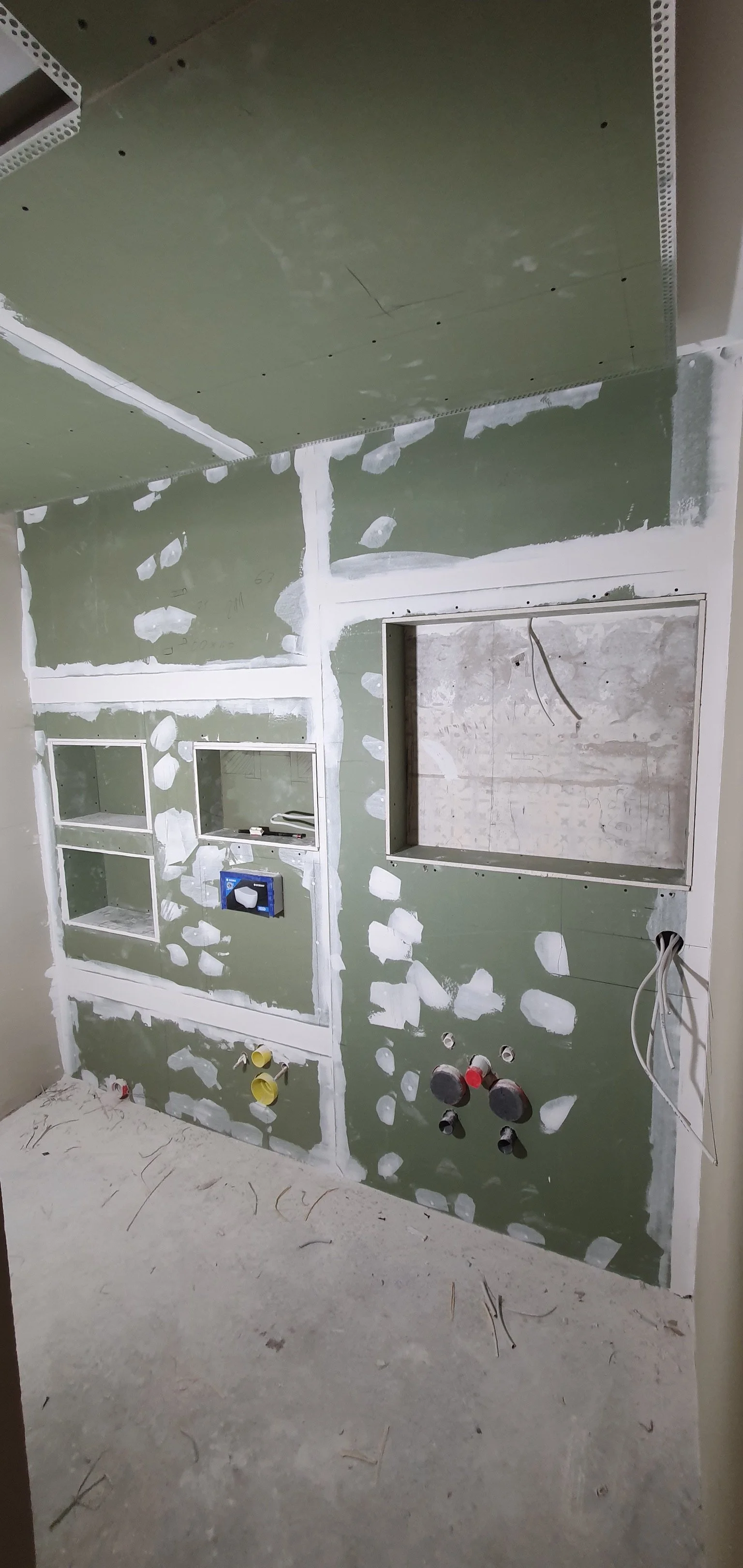 Raum under Renovierung mit grüner Trockenbauwand, offenen Nischen, Wasser- und Stromanschlüssen, auf dem Boden liegen Staub und Kabelreste.