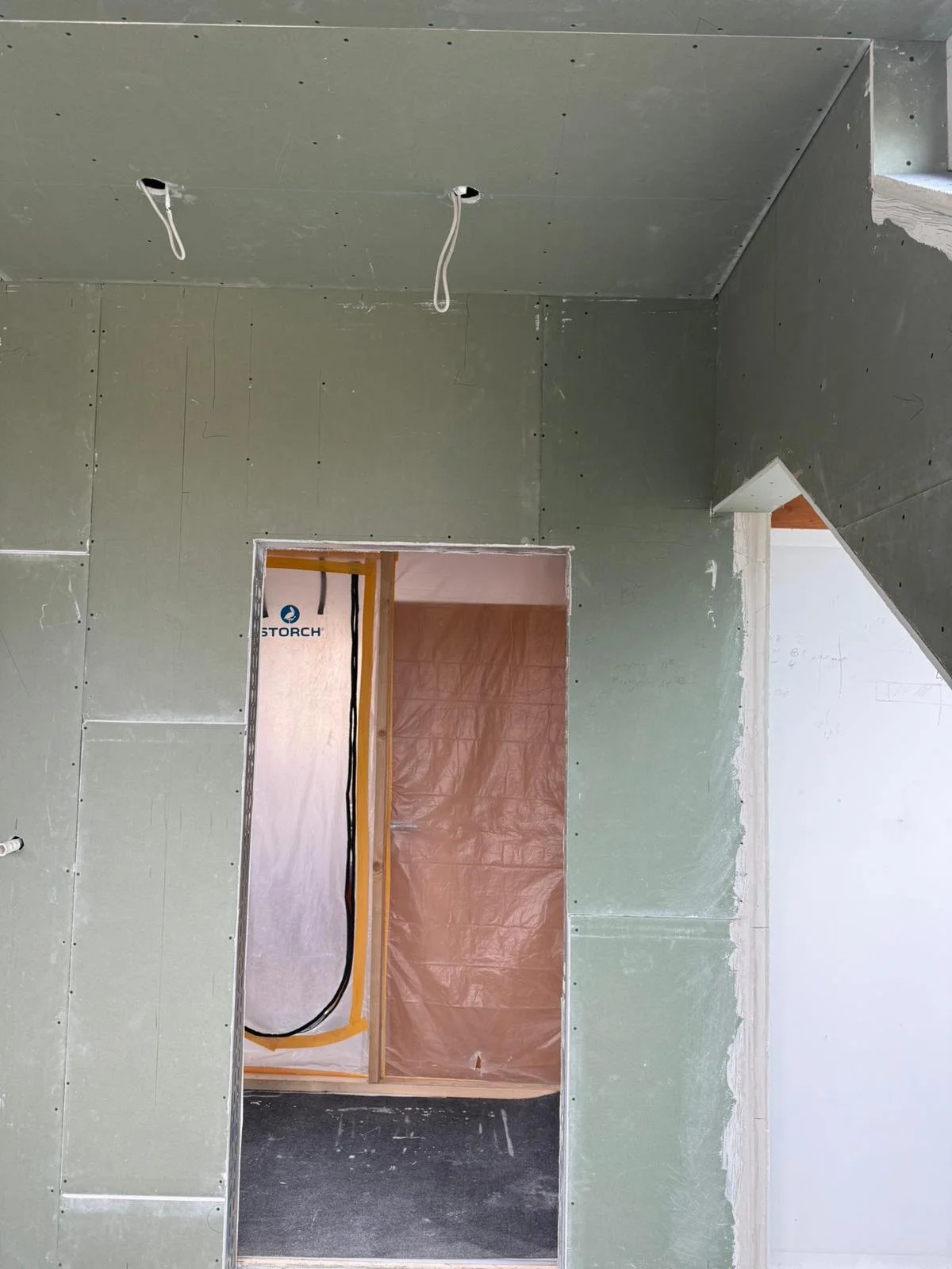 Innenansicht eines im Bau befindlichen Raumes mit grüner Trockenbauwand, zwei Kabel hängen von der Decke, Türrahmen ohne Tür, hinter dem Türrahmen sehen wir eine Kunststofffolie und eine weiße Wand.