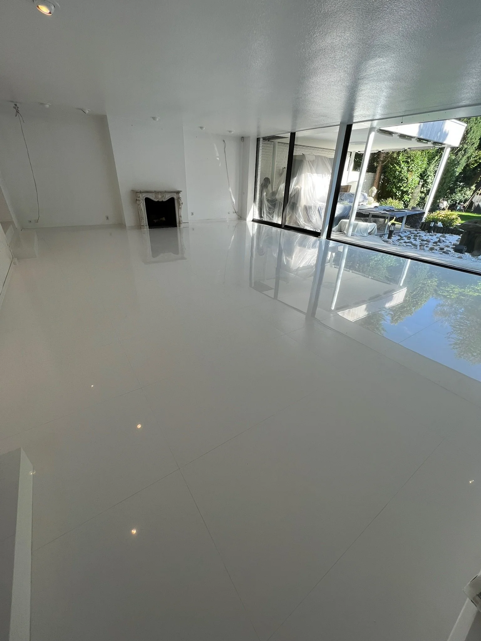 Leerer Raum mit weißem Boden, große Fensterfront und Kamin, draußen sichtbarer Garten mit Bäumen und Steinen.