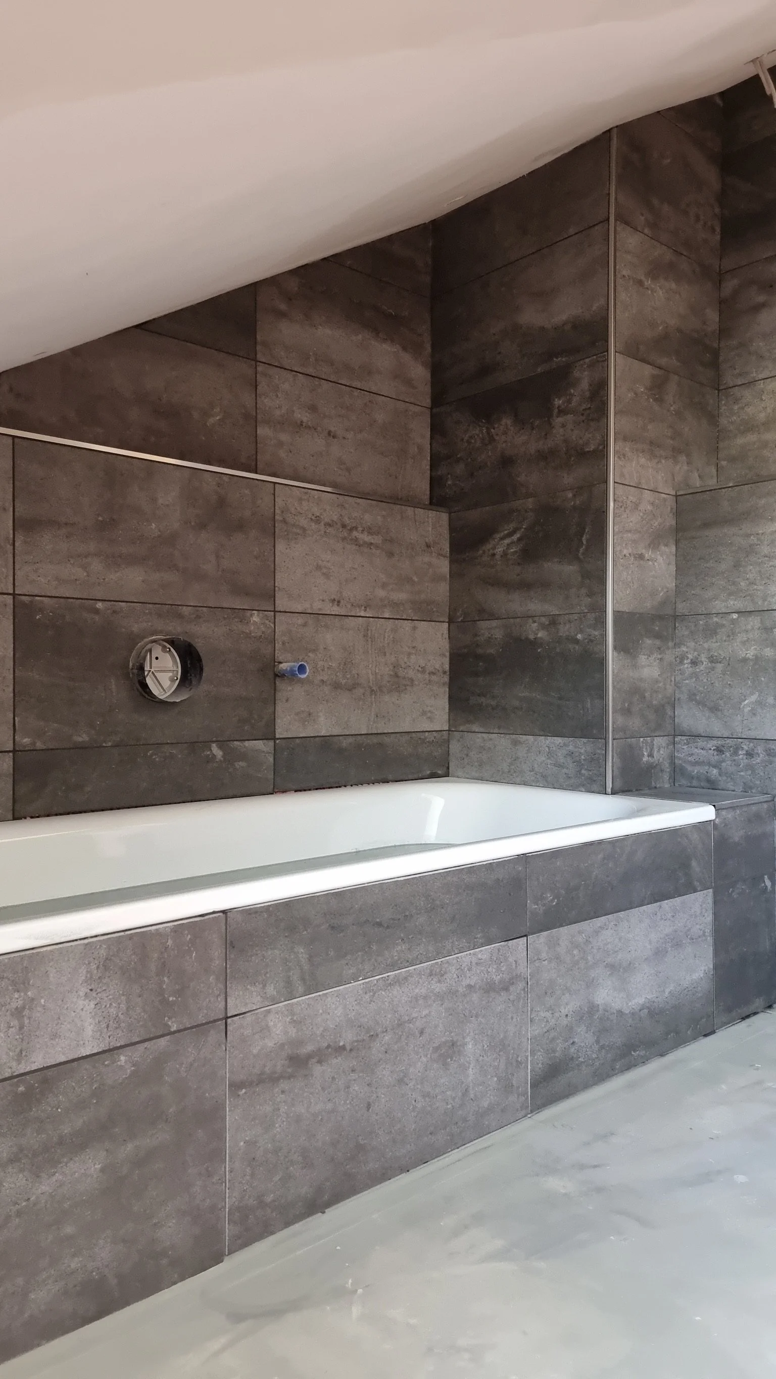 Ein Badezimmer mit dunklen Wandfliesen, einer Badewanne und einem unvollständigen Waschbeckenanschluss.