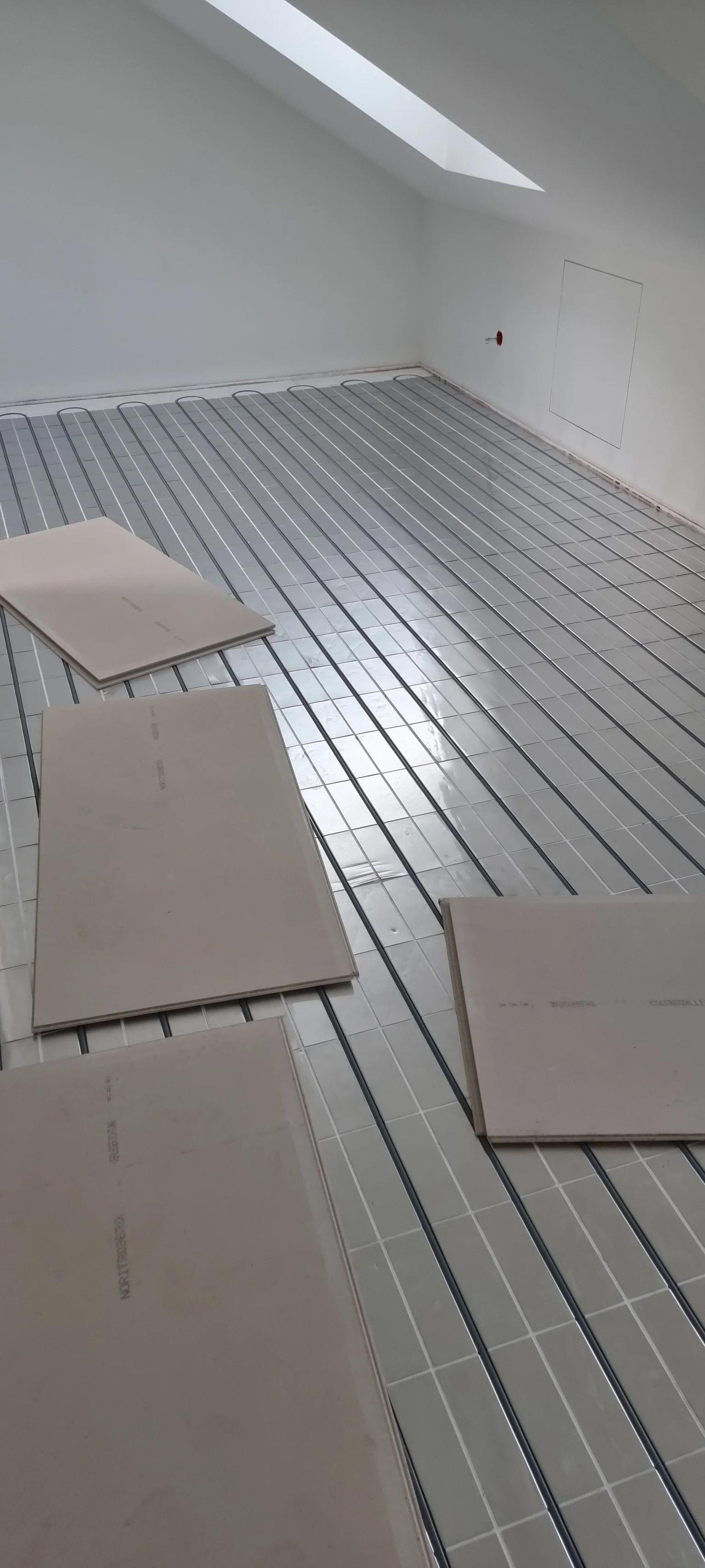 Ein Raum im Bau, der mit einer Fußbodenheizung ausgestattet wird, mit Heizrohren auf dem Boden; auf der linken Seite liegen Fliesen. Die Wände sind weiß, eine Dachschräge mit Fenster ist sichtbar.