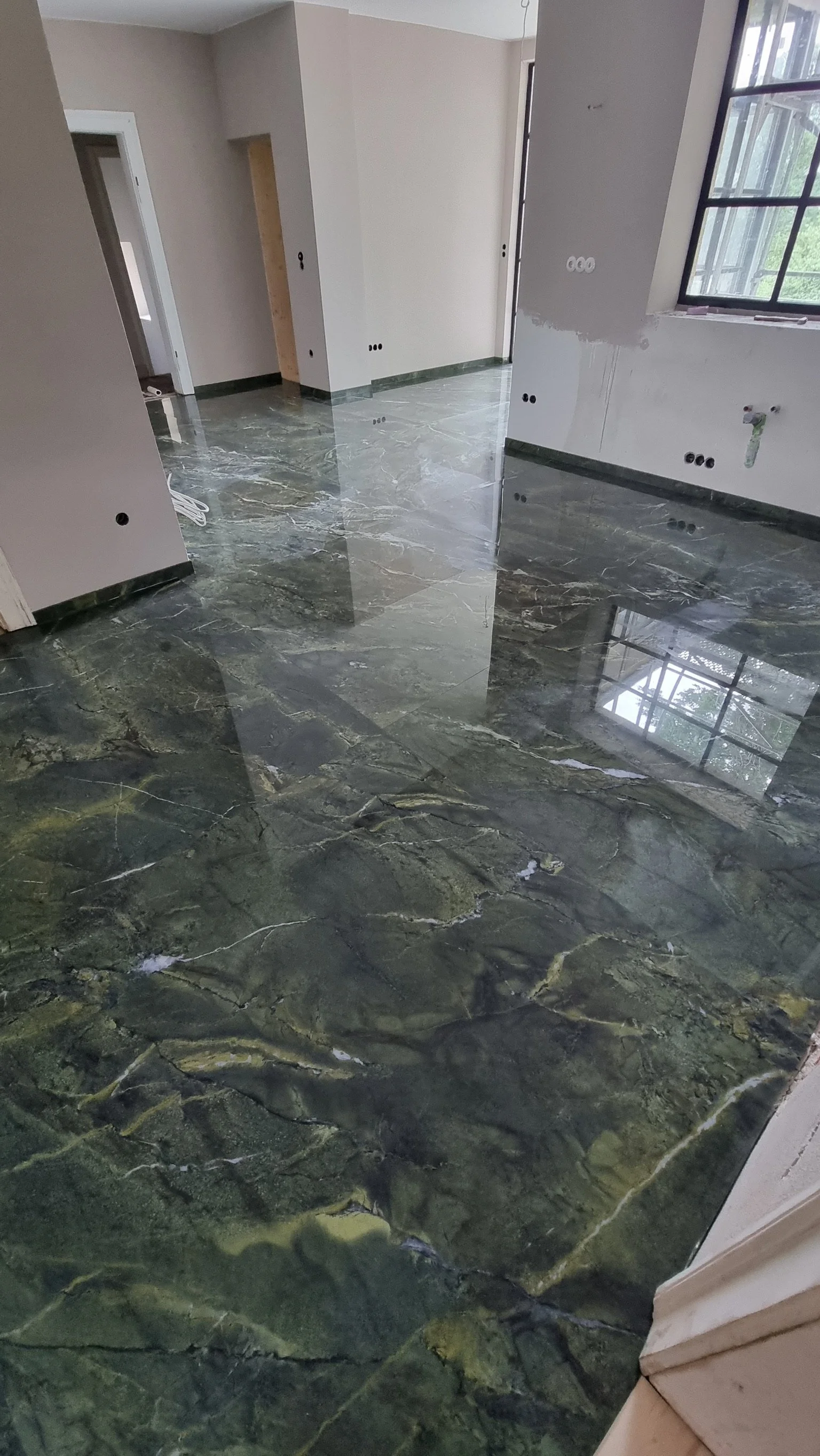 Innenansicht eines unfertigen Raumes mit poliertem, grün-schwarzem Marmorboden, weißen Wänden und großen Fenstern.