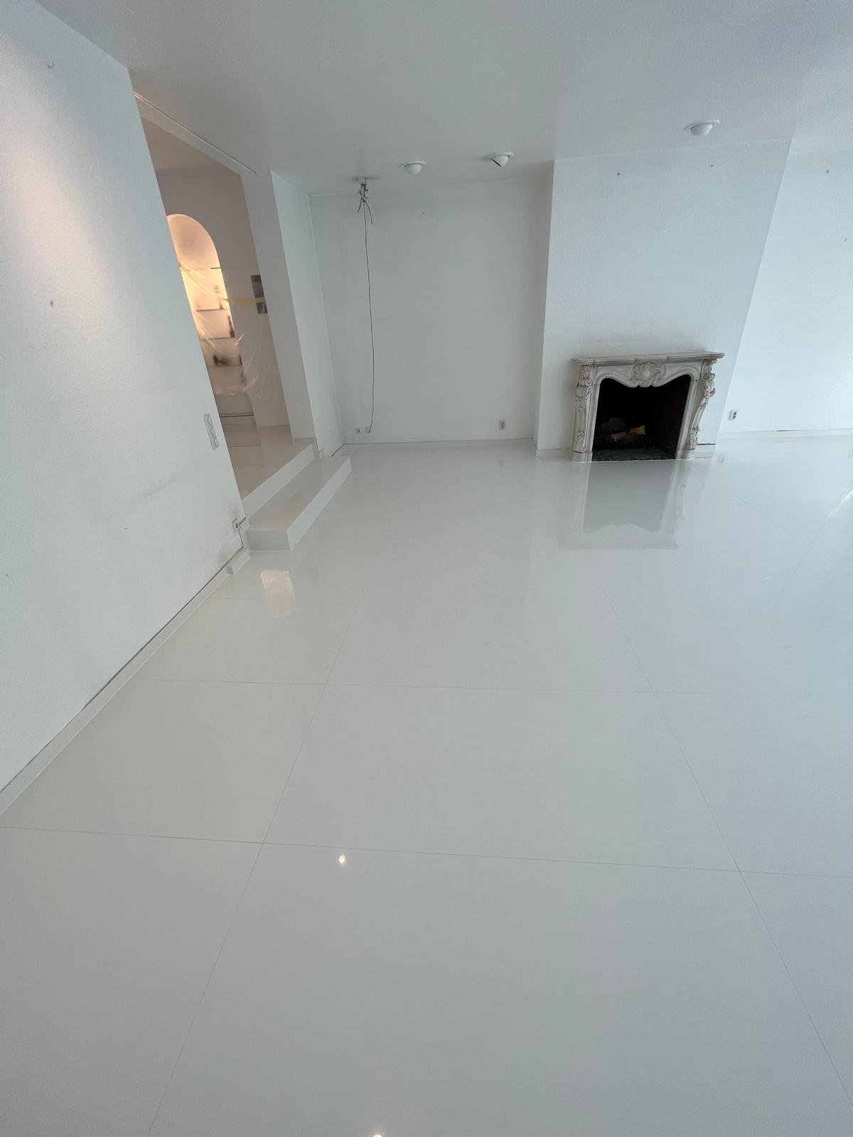 Ein leerer, weiß gestrichener Raum mit glatten weißen Fliesen, einer weißen Wand mit einem Kamin und einer offenen Tür im Hintergrund.