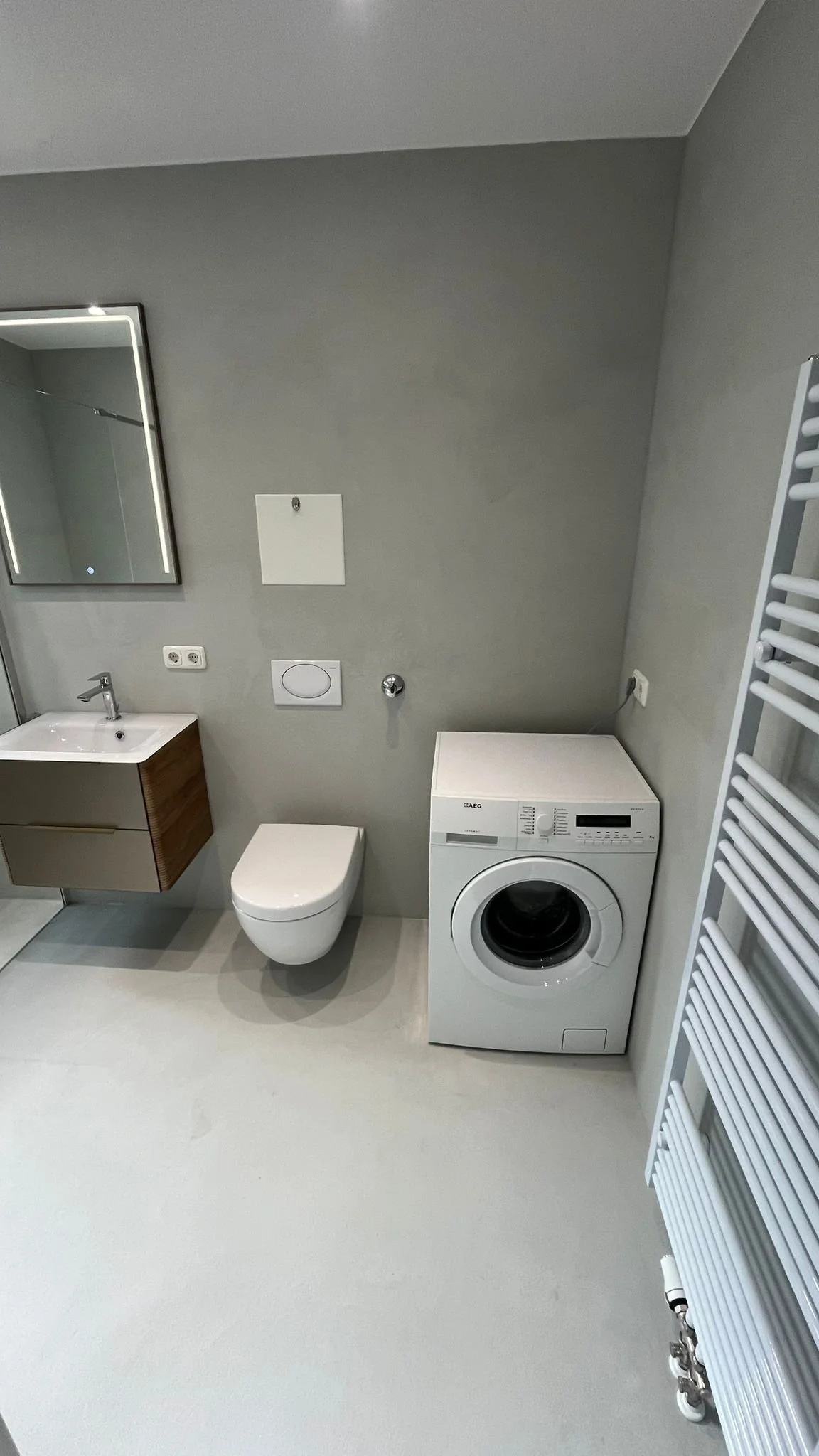 Ein modernes Badezimmer mit Waschbecken, Toilette, Waschmaschine, Spiegel, Föhn und Heizkörper.