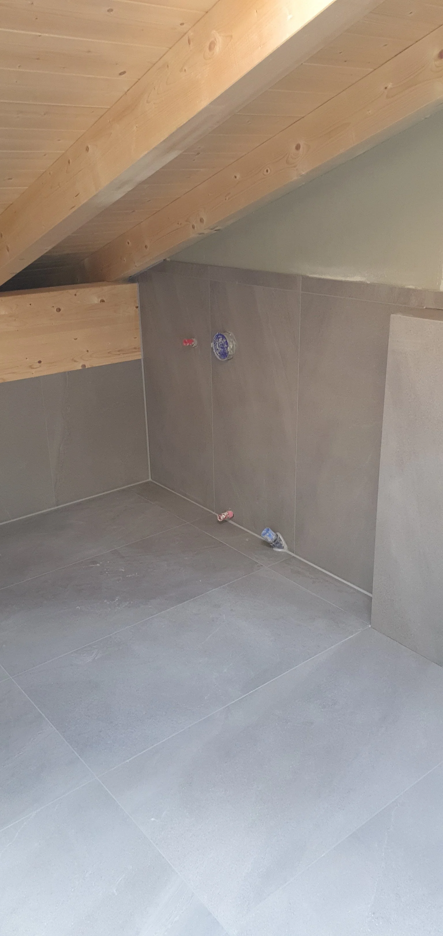 Innenraum unter einem Dach mit Holzdecke und Tapetenwand, unbenutzte Wasseranschlüsse und Abflussrohre, Boden und Wände mit Fliesen