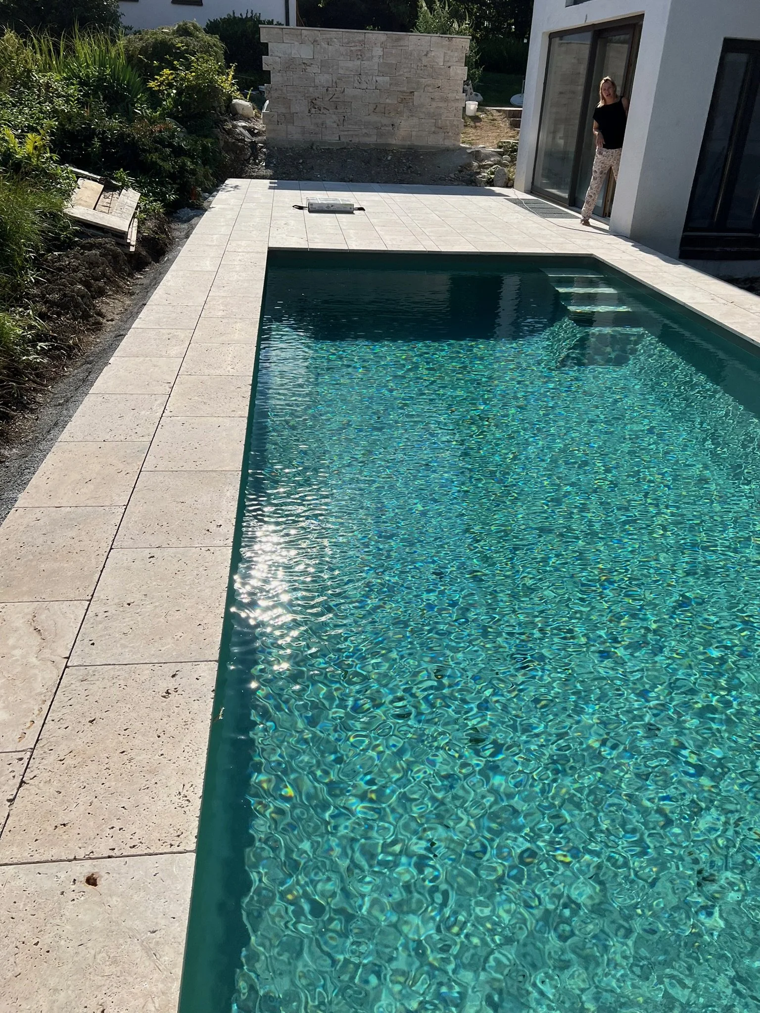 Ein moderner Swimmingpool mit Wasser in einem Garten neben einem weißen Haus, eine Frau steht am Rand des Pools.