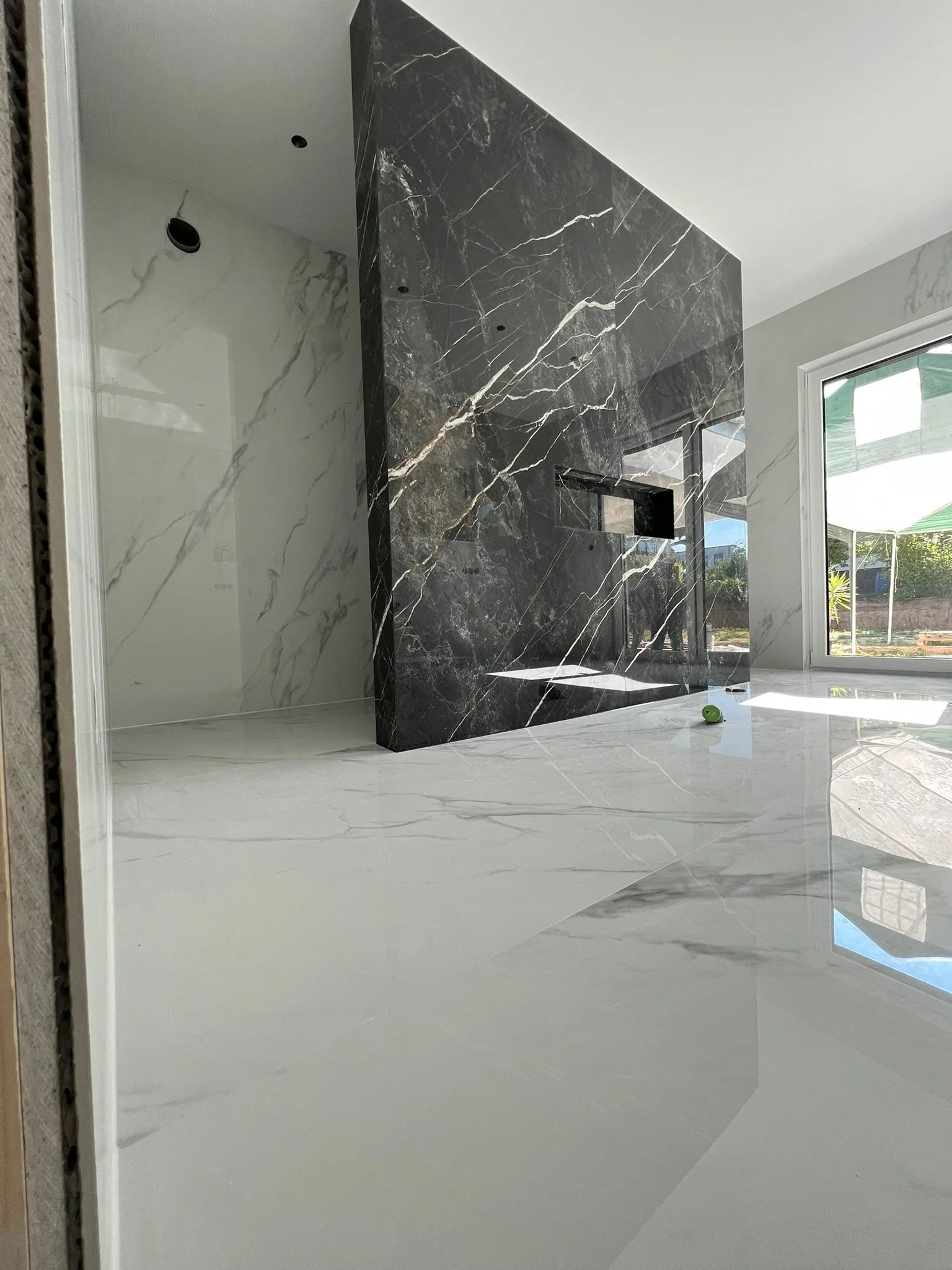 Ein moderner, heller Raum mit großen weißen Marmorfliesen auf dem Boden und einer schwarzen Marmorwand mit integtriertem Kamin. Große Fenster lassen viel Tageslicht herein und bieten einen Blick nach draußen.