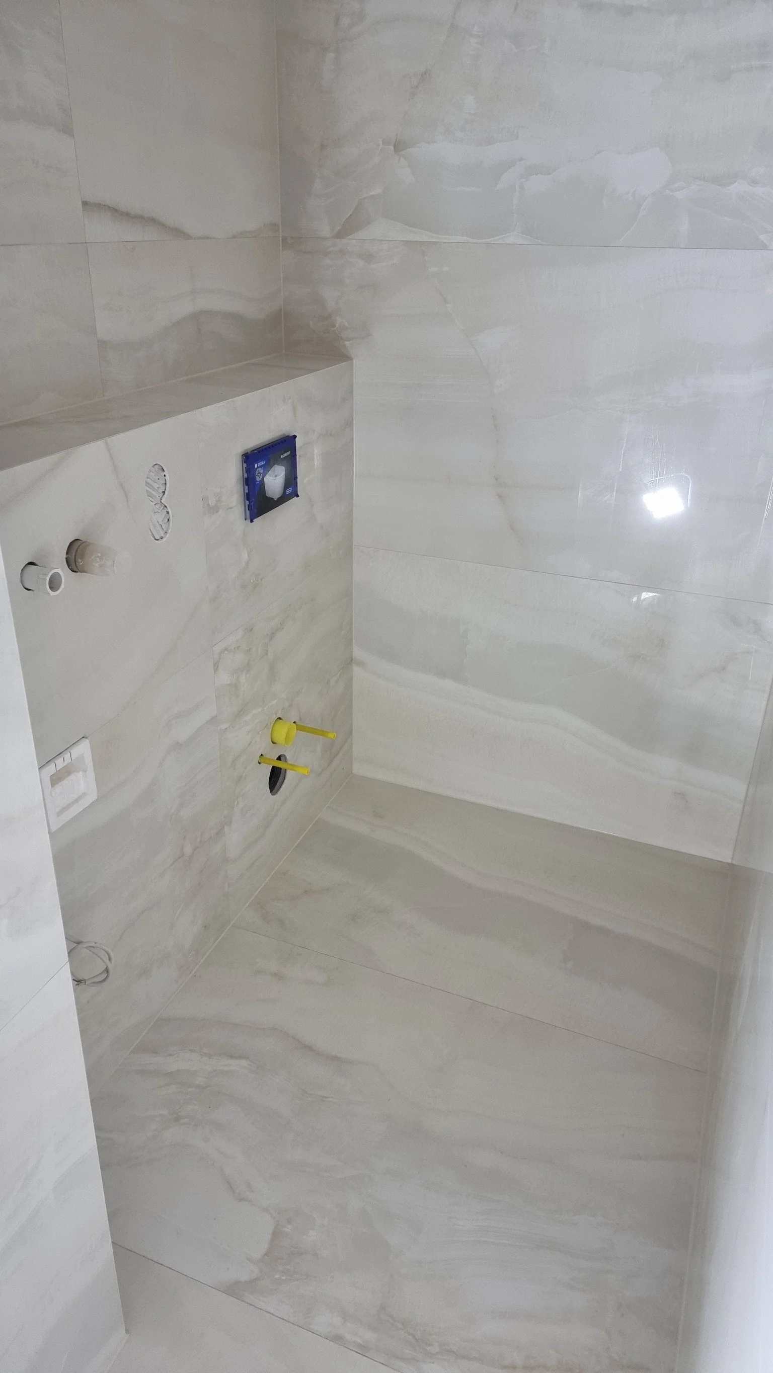Ein unfertiges Badezimmer mit Wand- und Bodenfliesen aus hellem Marmor, installierten Wasseranschlüssen und einem versteckten WC.