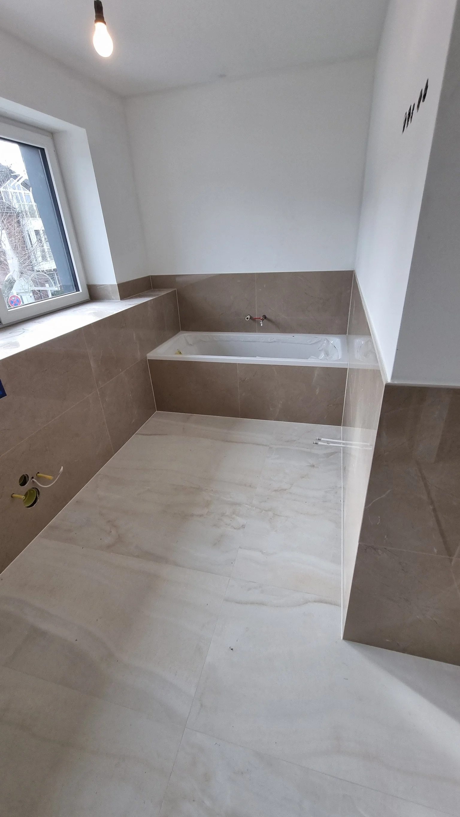 Ein Badezimmer unter Bau, mit einer Badewanne, einem Fenster und unvollständiger Wasser- und Strominstallation.