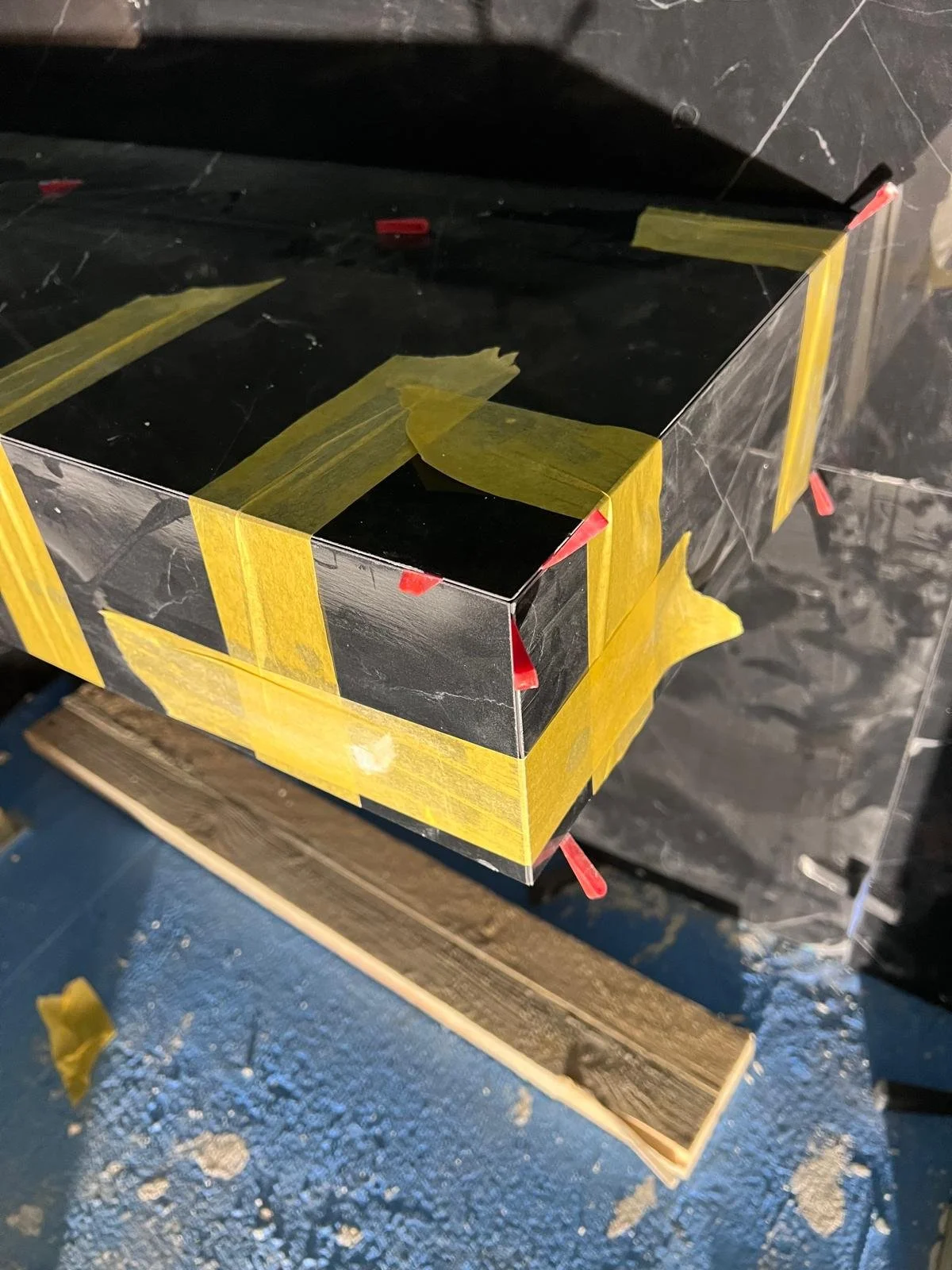 Ein schwarzer, spiegelnder Würfel, in dem sich die Umgebung spiegelt, mit gelbem Klebeband an den Kanten. Der Würfel steht auf einem Stück Holz auf einer blauen Oberfläche, mit Staub und Spänen. 