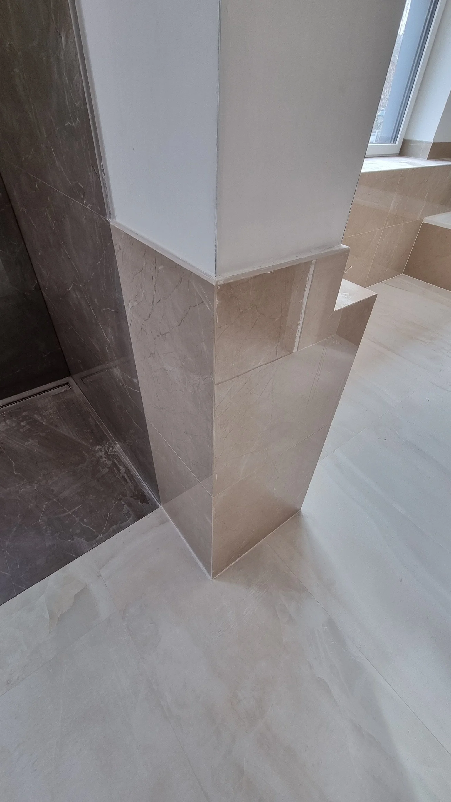 Ecke eines Badezimmers mit Wandfliesen in Beige, weißem Putz, dunklen und hellen Fliesen, und einer Fensterbank.