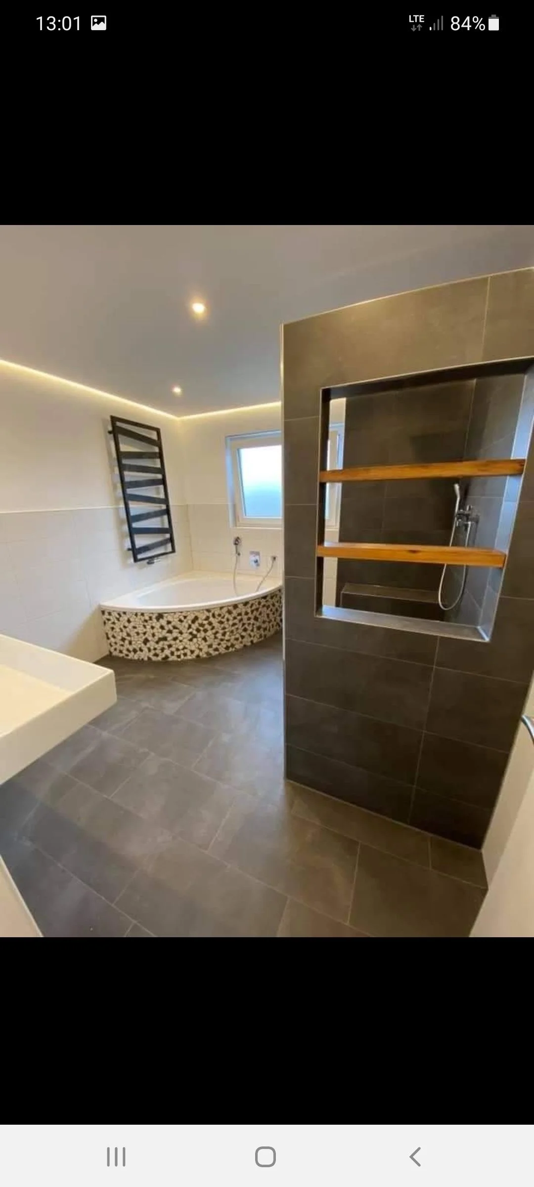 Modernes Badezimmer mit dunklen Fliesen, einer Ecke mit Badewanne, schwarzem Handtuchhalter und einer Duschnische mit Holzregalen.