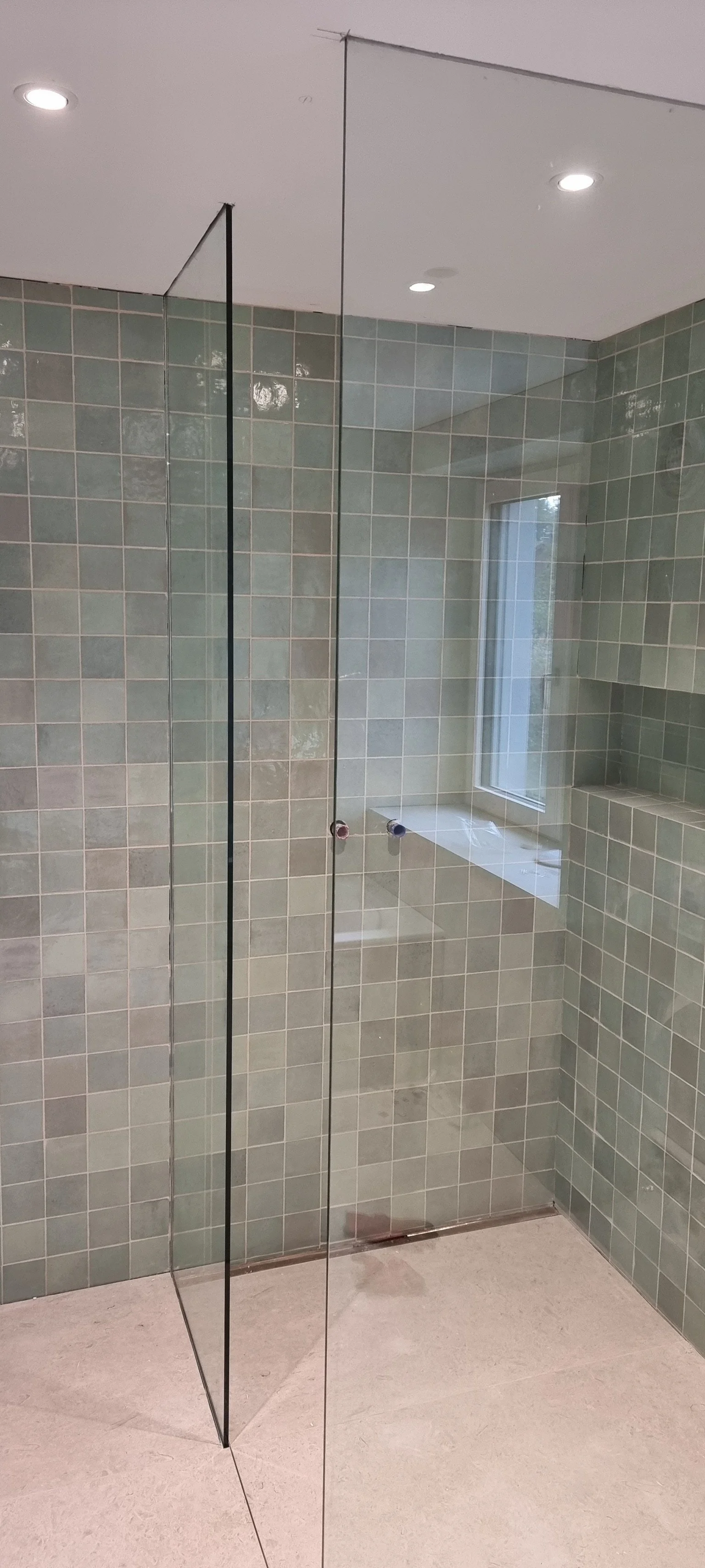 Ein moderner, unfertiger Duschbereich mit grünlich glasierten Fliesen, einem großen Fenster, einer Glastür und integrierten Ablagefächern.