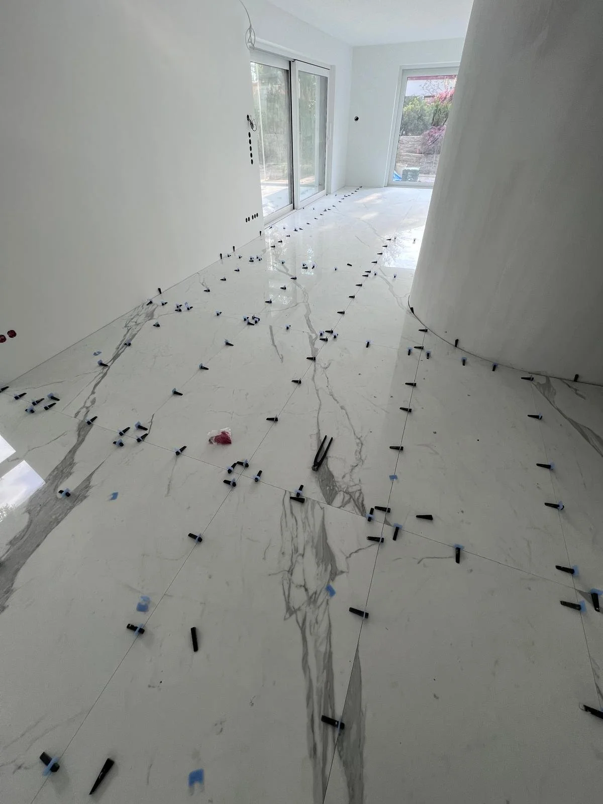 Innenraum eines Hauses im Bau, mit großen weißen Fliesen auf dem Boden, die noch verlegt werden, inklusive Verlegezubehör und Fliesenmörtel, sowie große Fenster mit Blick ins Freie.