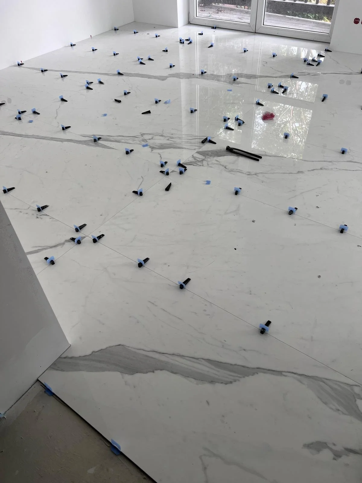 Ein Raum mit einer weißen Marmor-Dielung, auf der Fliesenverlegeklemmen verteilt sind, um die Fliesen während der Verlegung zu fixieren. Es gibt große Fenster, die Tageslicht hereinlassen.