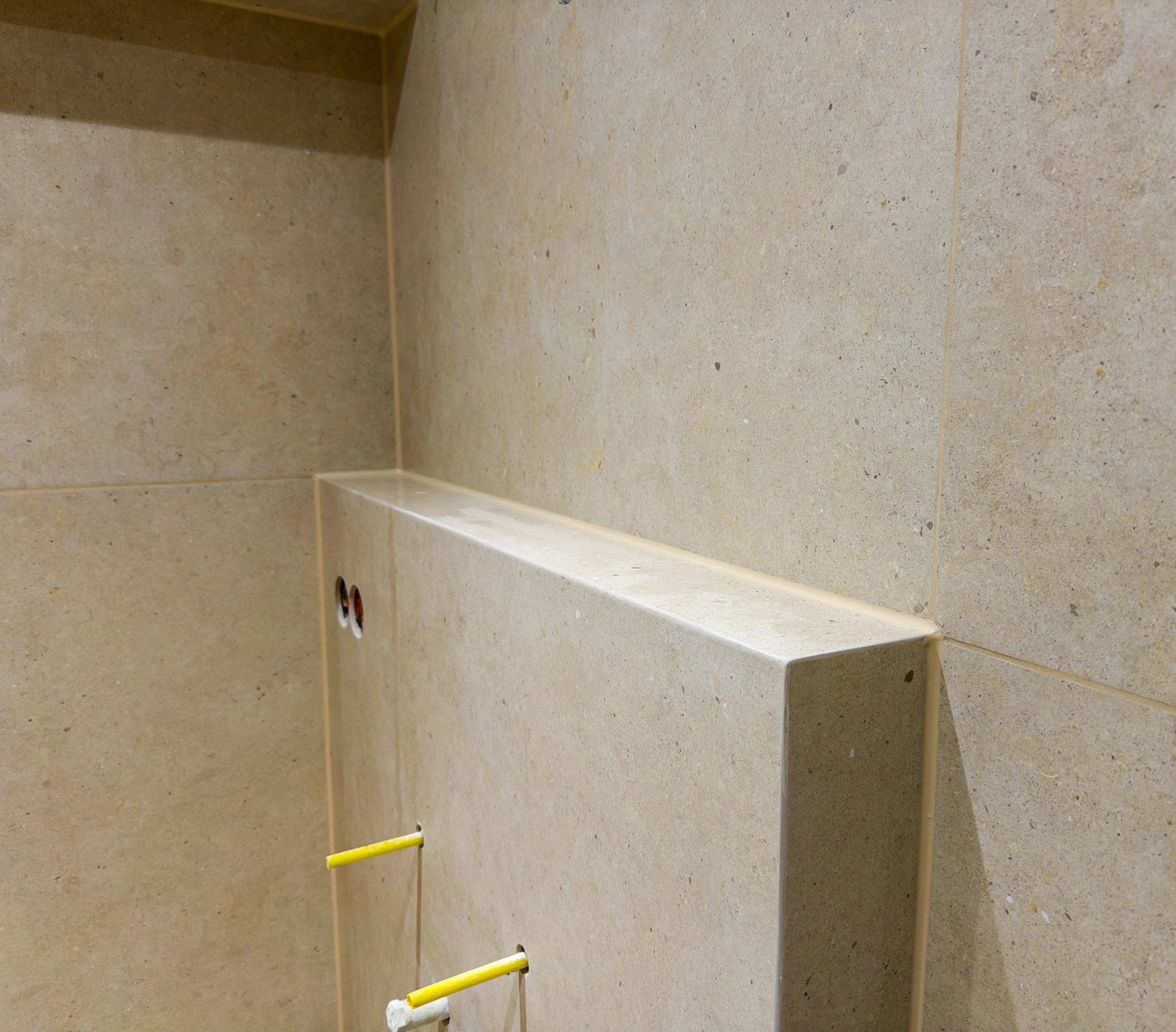 Innenaufnahme einer Wand mit beige Fliesen, eine hervorstehende schmale Ablage aus dem gleichen Material, zwei kleine Löcher und zwei gelbe Rohrleitungen.