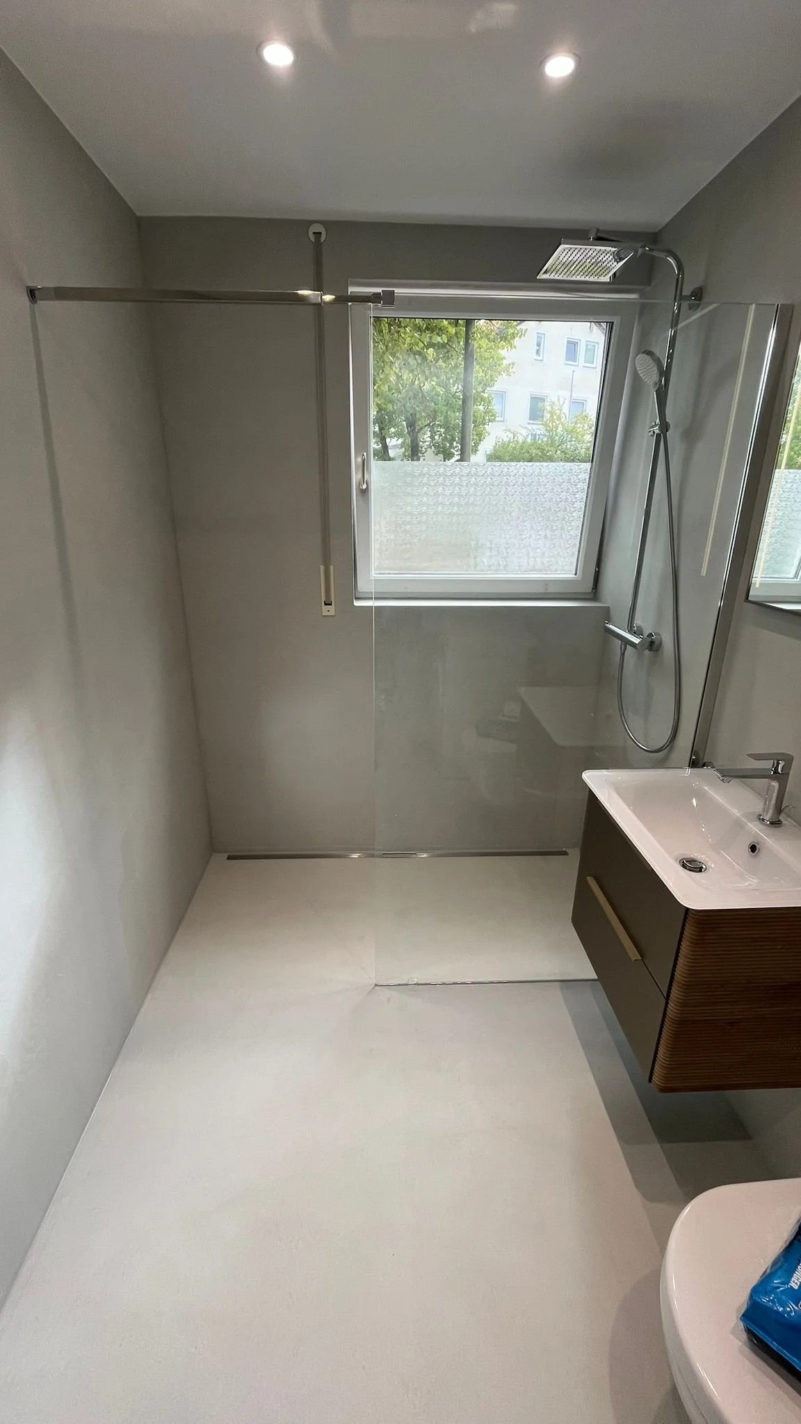 Ein modernes Badezimmer mit begehbarer Dusche, weißem Boden, grauen Wänden, einem Fenster mit Blick auf Bäume, einem weißen Waschtisch mit dunklem Unterschrank und einer Halterung für eine Duschstange.