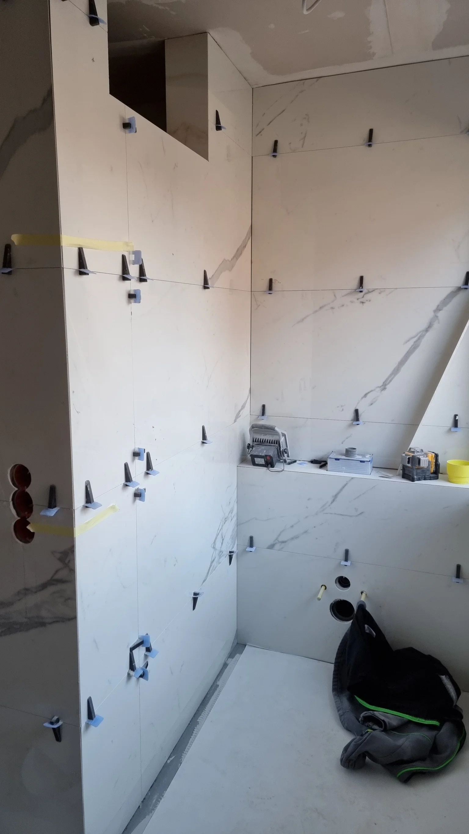 Zimmer im Bau mit weißen Wandfliesen und schwarzen Fliesenklemmen, Werkzeuge auf einer Ablage, eine schwarze Tasche im Vordergrund.