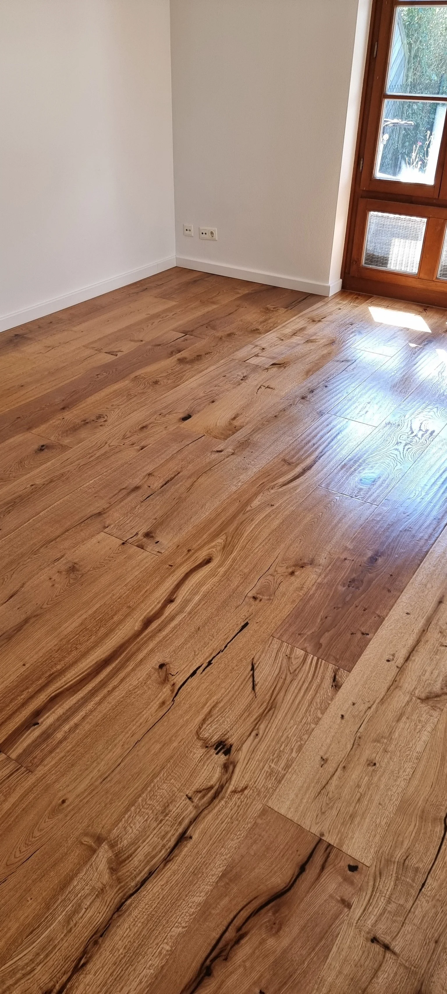 Ein heller Raum mit Holzboden, weiße Wände, ein Fenster und eine Steckdose an der Wand.