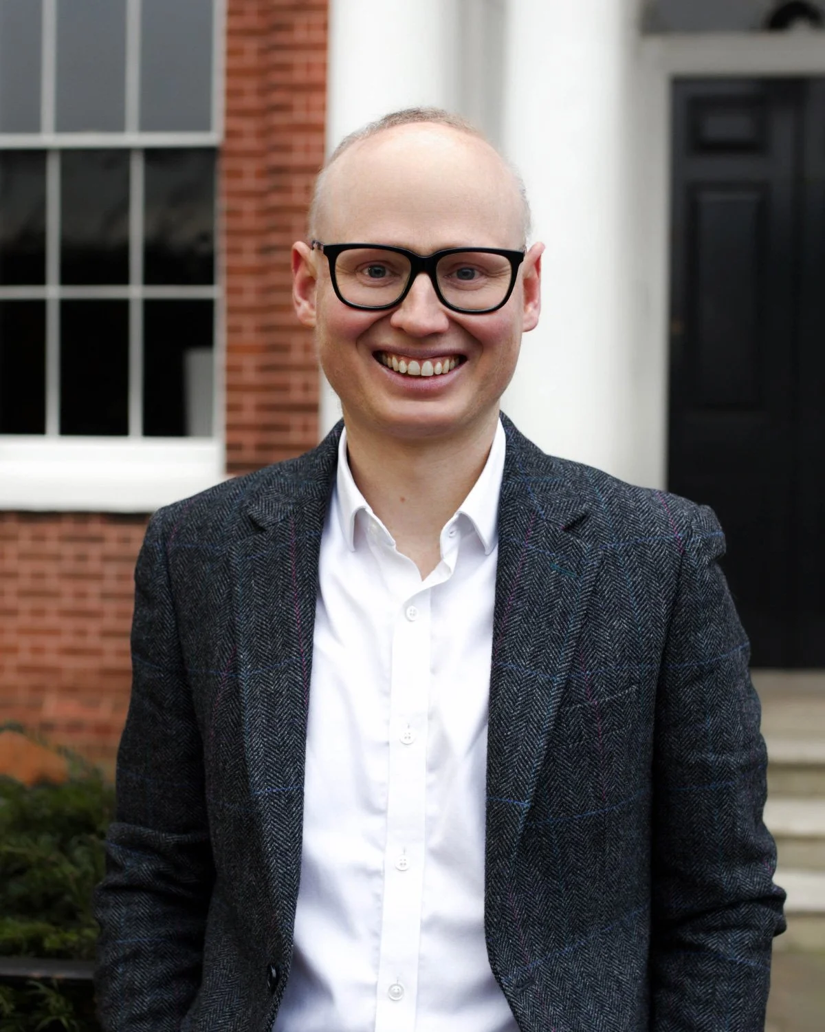 Man wearing glasses and a light-colored shirt, smiling with a hand on his chin, against a blue background.